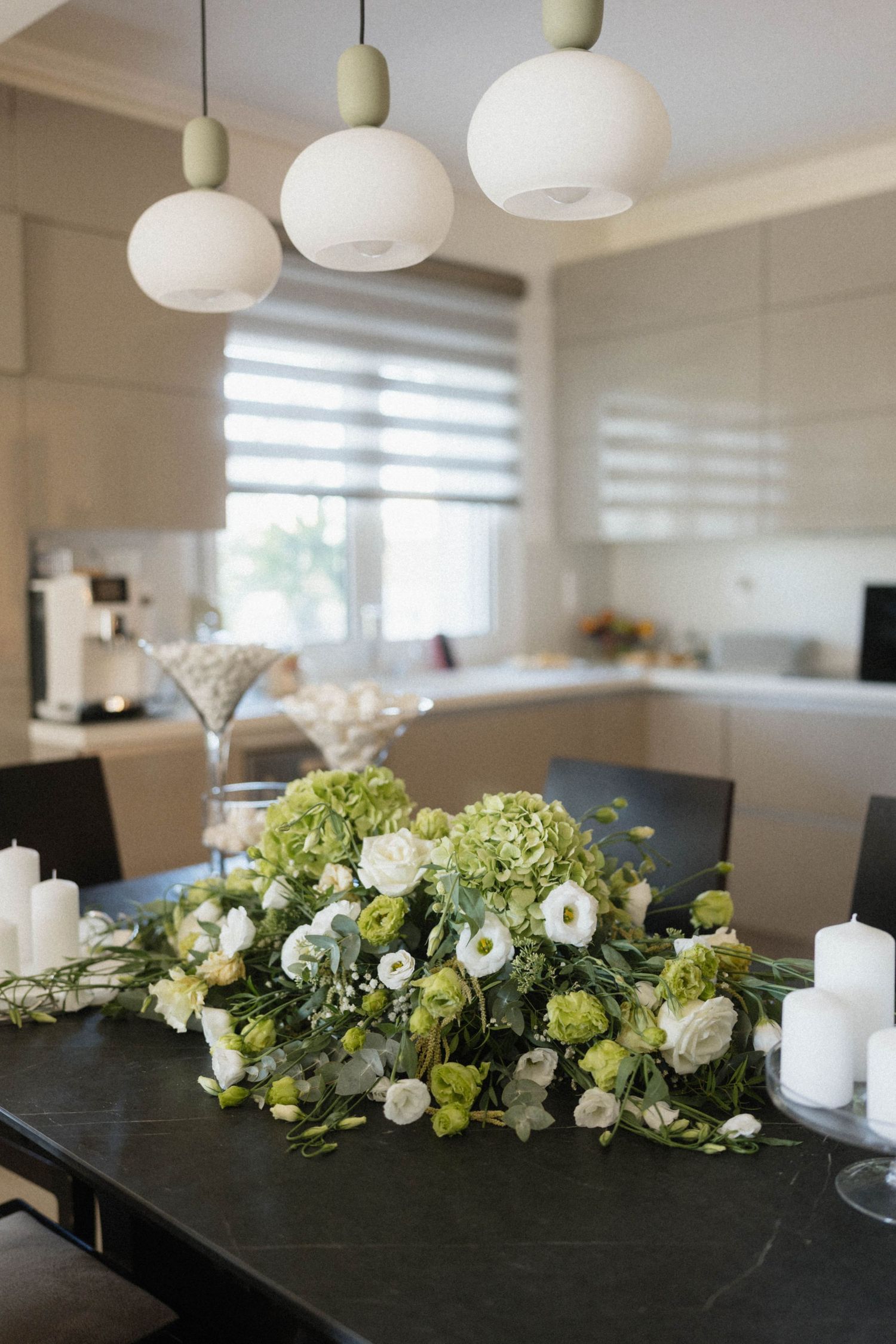 Elegant dining table with green and white floral centerpiece, white pillar candles, and globe pendant lights in modern kitchen.