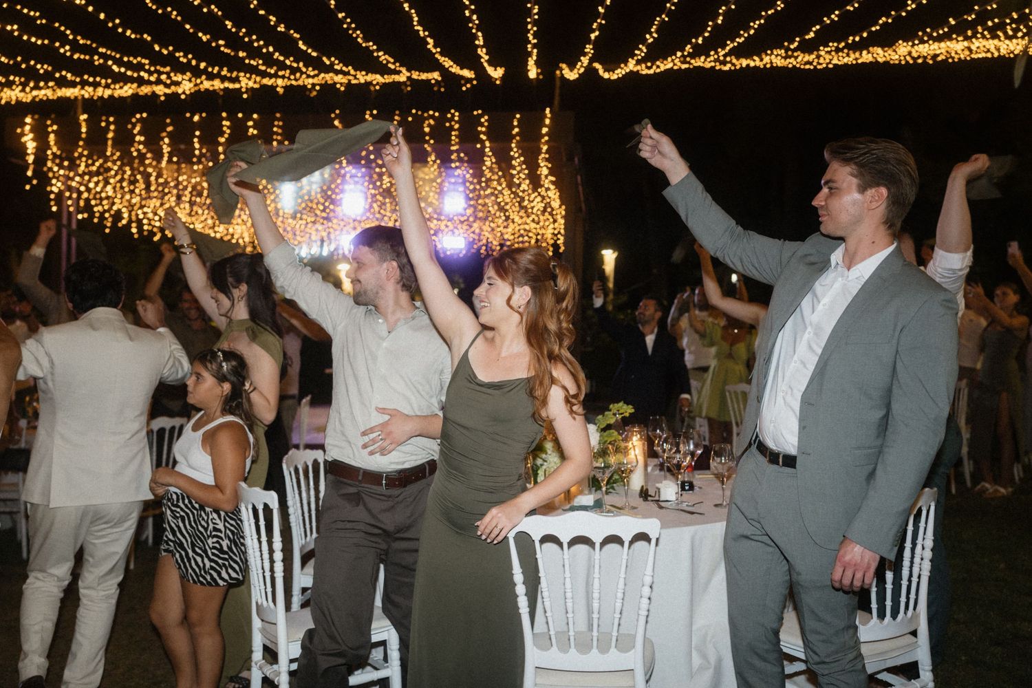Guests dancing and celebrating at an outdoor wedding reception under warm string lights at night.