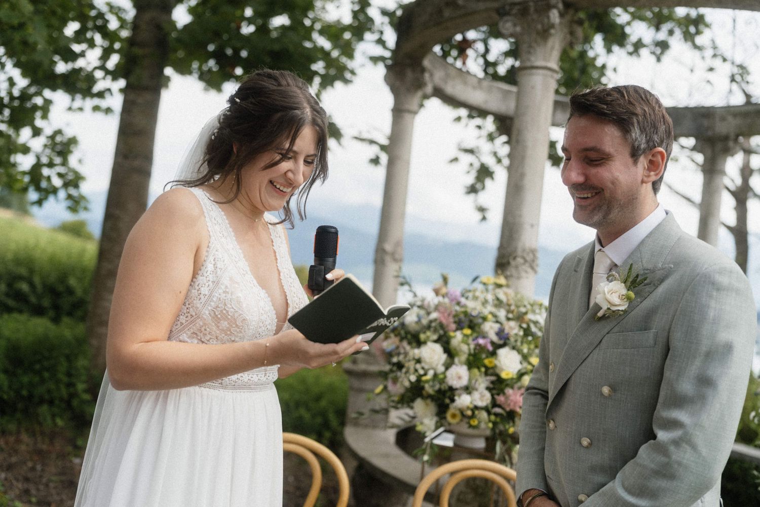 Bride reading vows to smiling groom at outdoor wedding ceremony with floral arrangements.