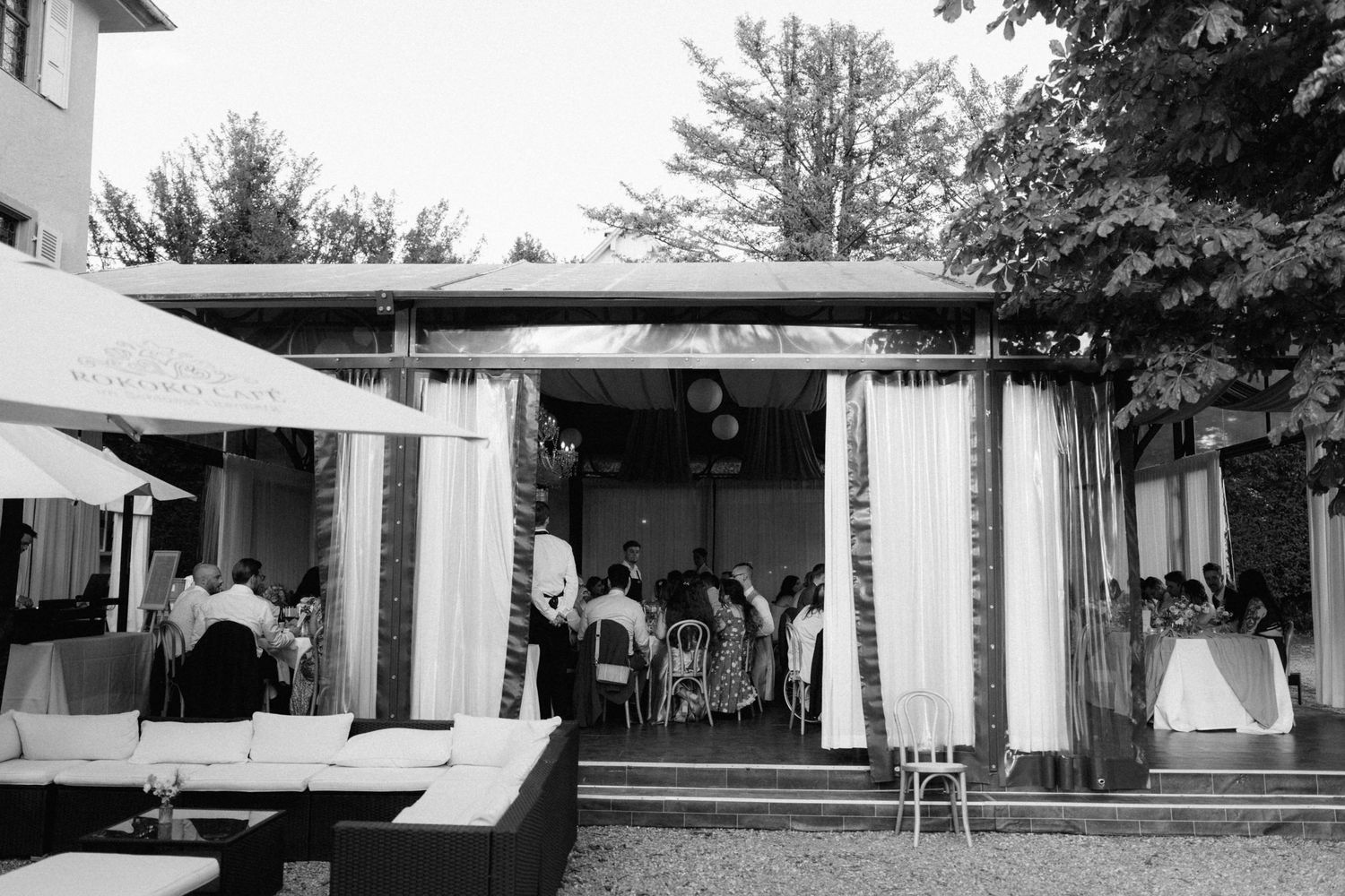 Black and white photo of wedding guests gathering at a draped outdoor reception pavilion at dusk.