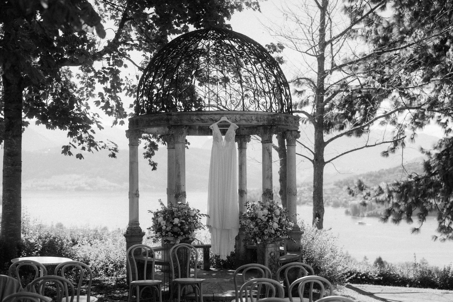 Black and white photo of an elegant outdoor wedding ceremony setup under a stone gazebo overlooking a serene lake.