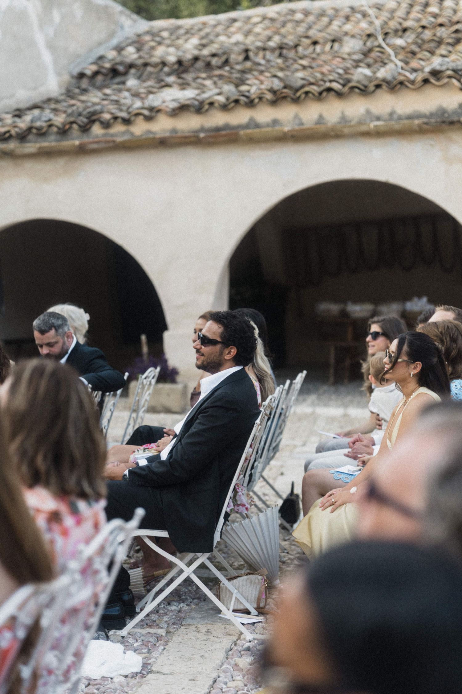 Wedding guests seated in ornate chairs at outdoor ceremony beneath stone arched building.