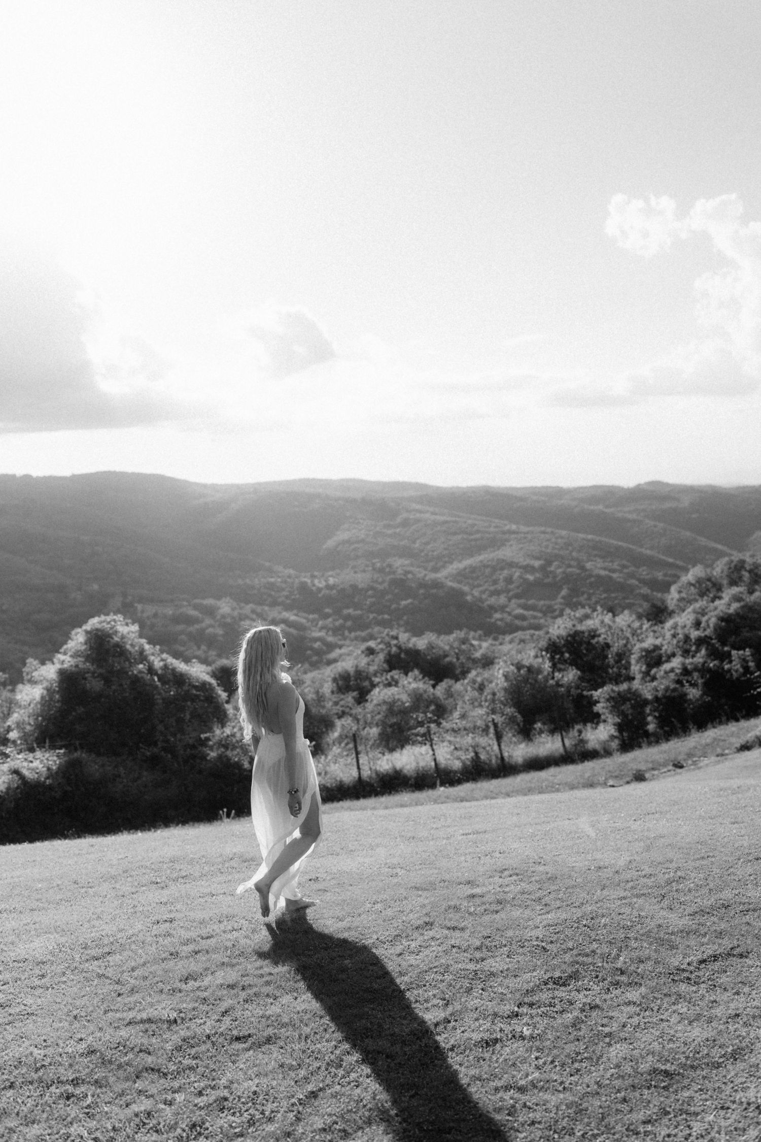 Black and white photo of a woman in a dress walking on a hillside meadow with scenic mountain views.
