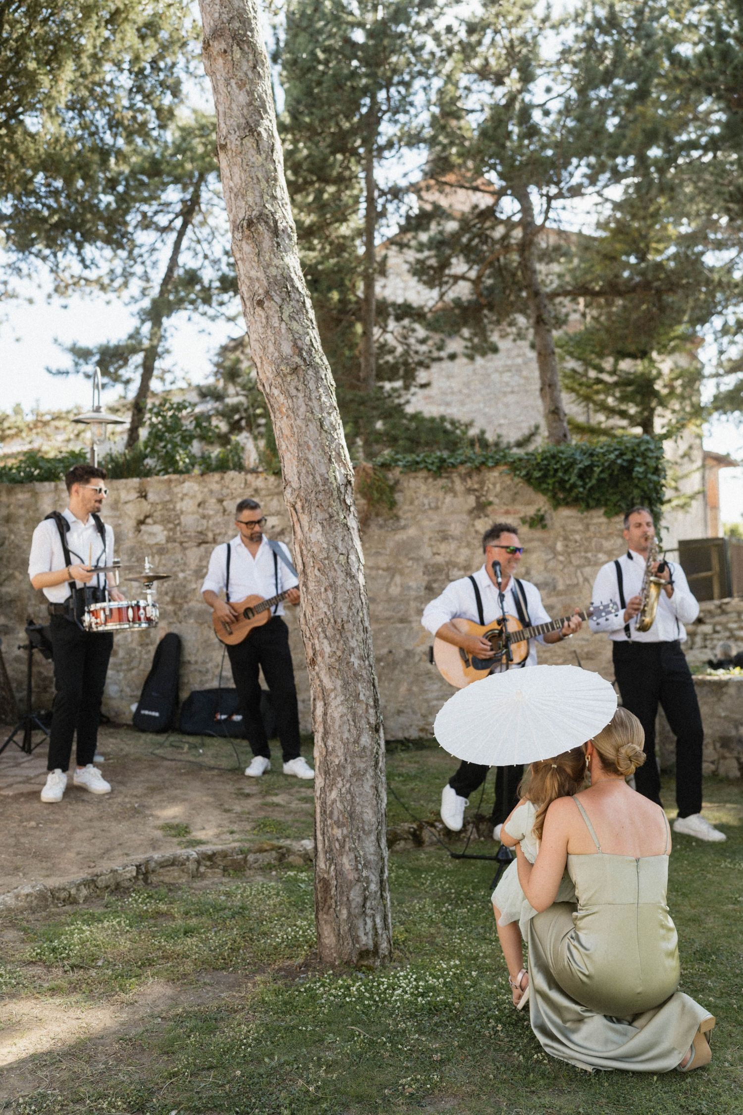 Live band performing outdoors at a wedding while a guest watches under a white parasol.