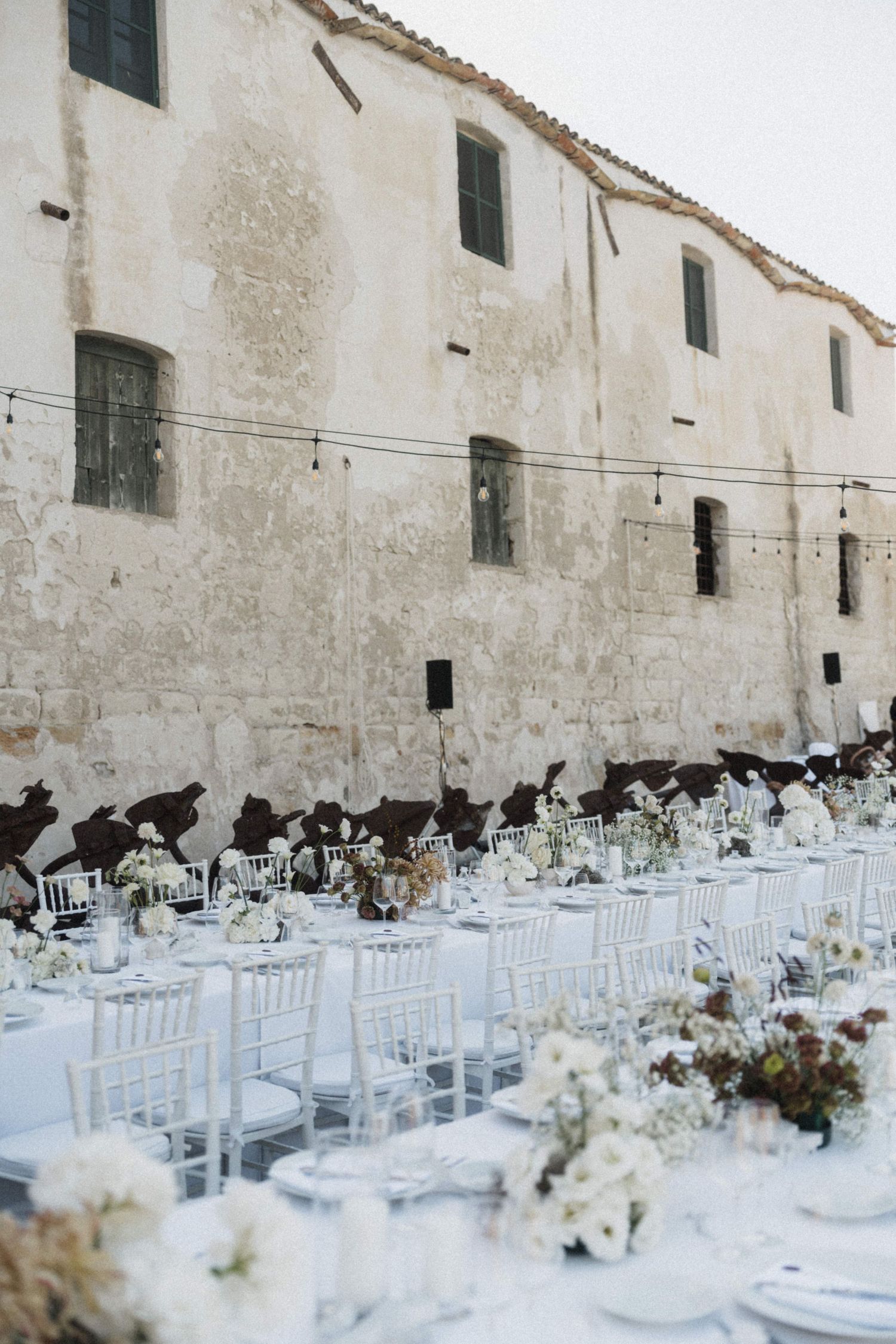 Outdoor wedding reception with long white tables set against a rustic stone building facade.