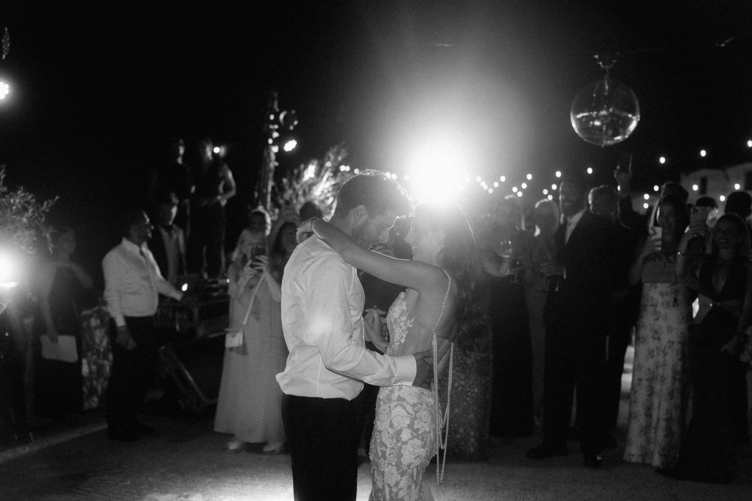 Black and white photo of bride and groom sharing first dance surrounded by guests at nighttime wedding.