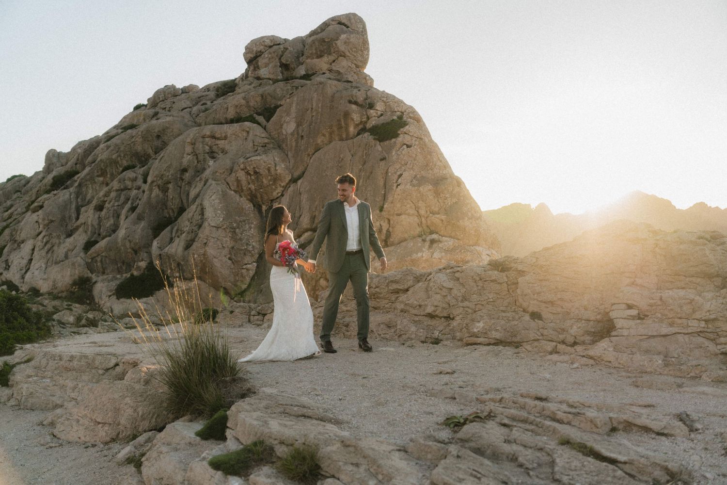 Bride and groom hold hands on rocky desert landscape at sunset near large boulder formation.