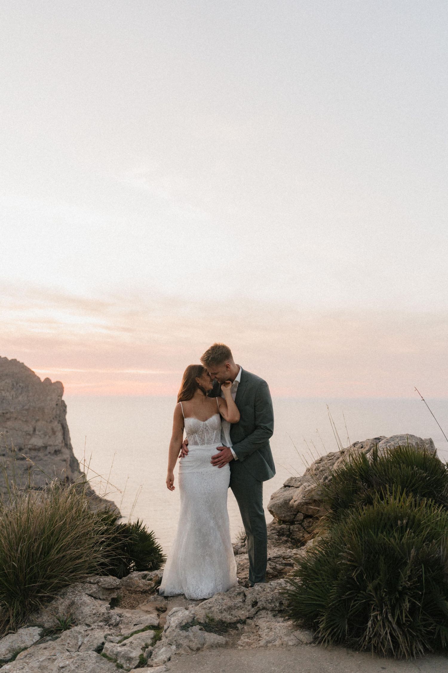 Bride and groom share a tender moment on rocky coastal cliffs at pastel pink sunset by the sea.