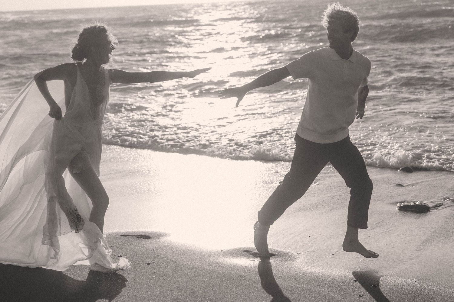 Black and white photo of couple dancing playfully on sunlit beach shoreline.