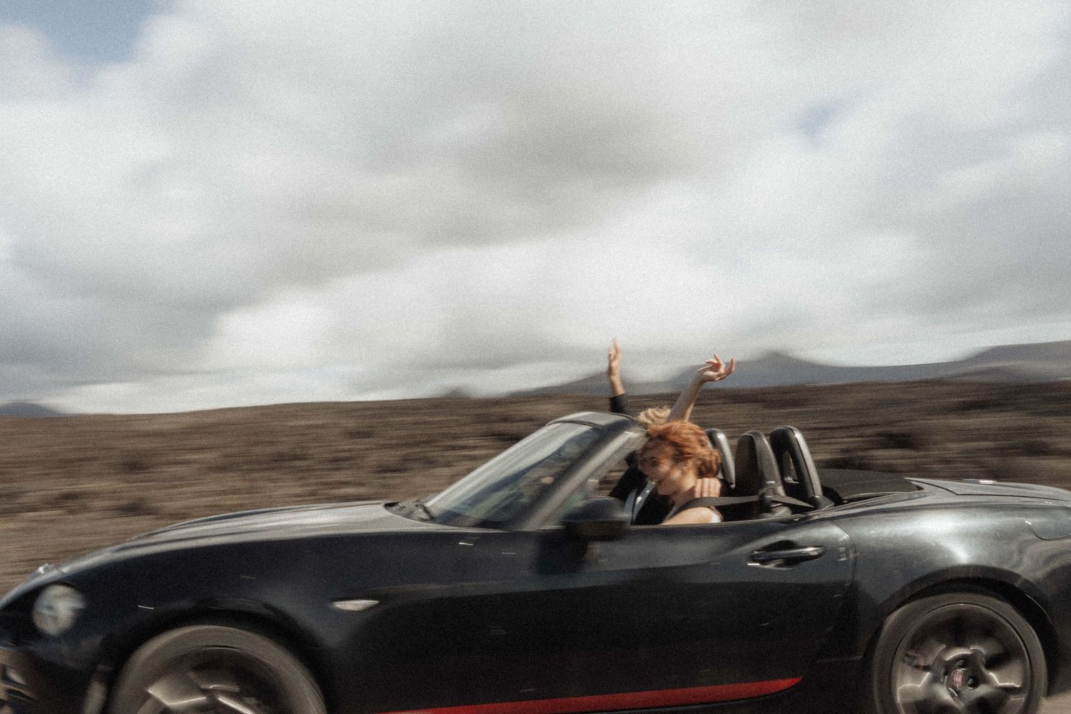 Woman with red hair riding in a black convertible sports car with hands raised, driving through open landscape.