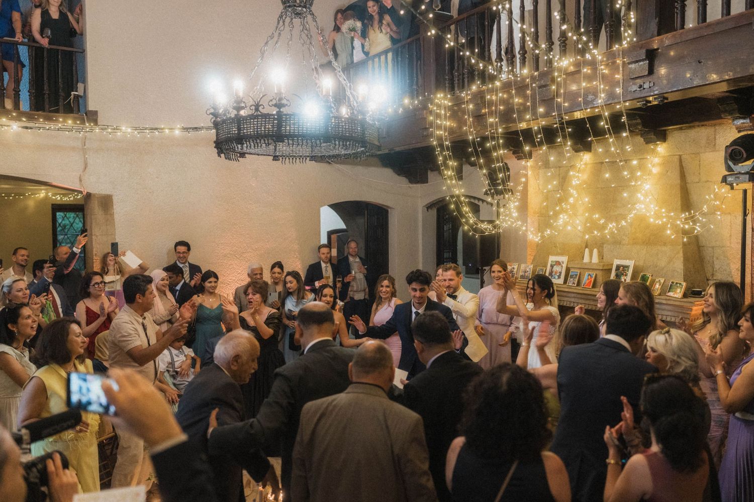 Wedding guests celebrate on dance floor inside rustic venue decorated with warm fairy lights.