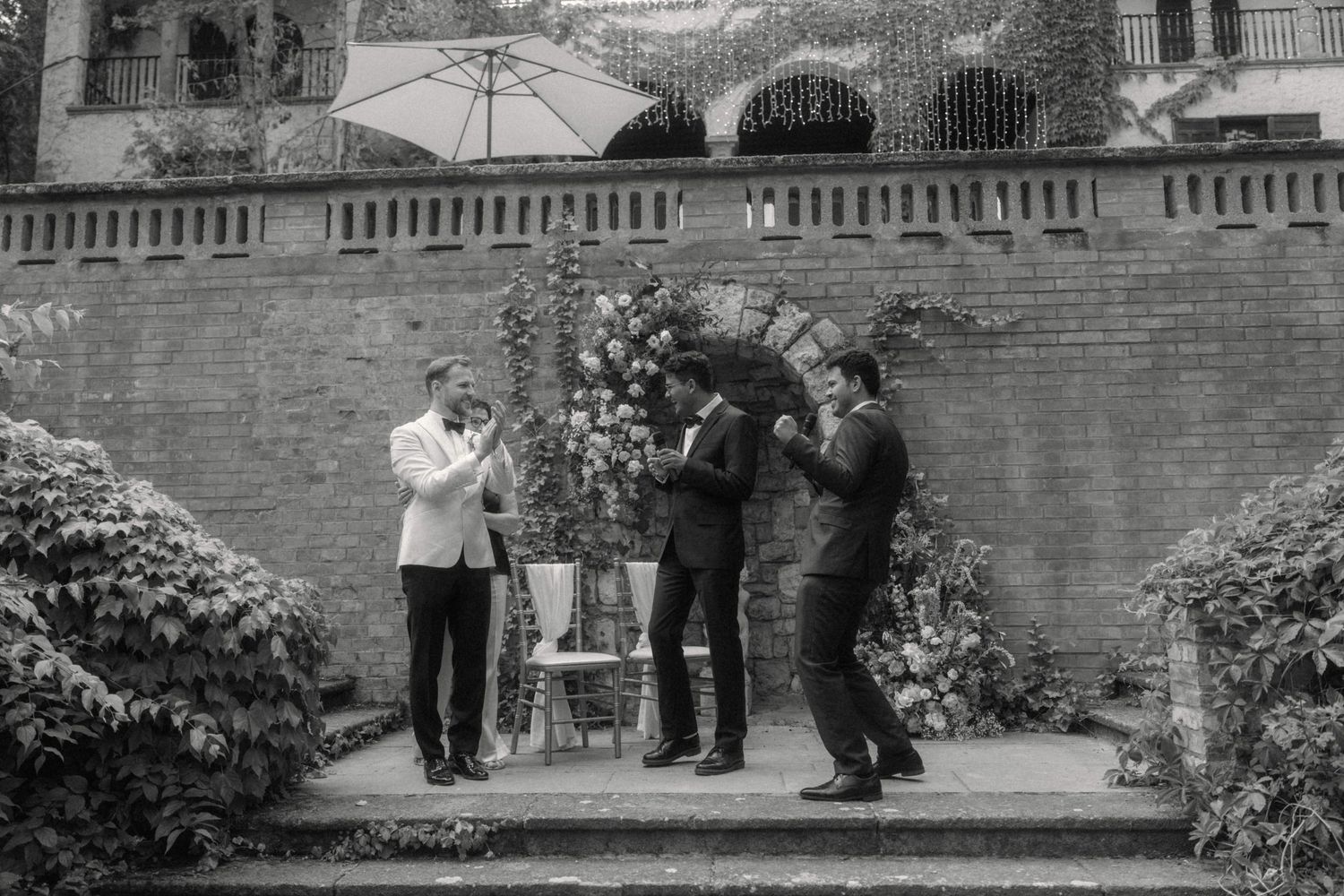 Black and white photo of three men in suits gathered outdoors at a wedding ceremony.