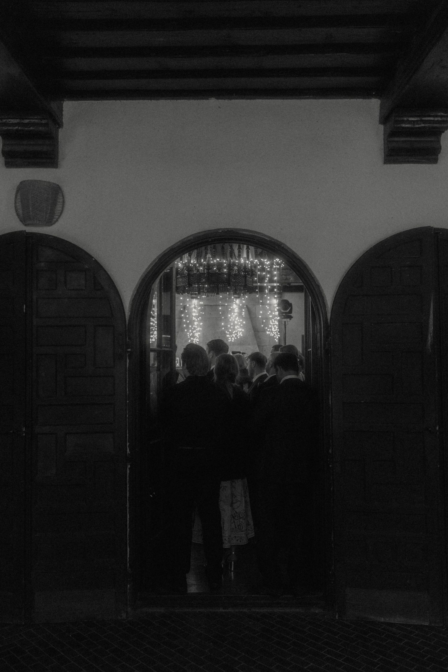 Black and white photo of wedding guests silhouetted in an arched doorway with sparkling lights inside.