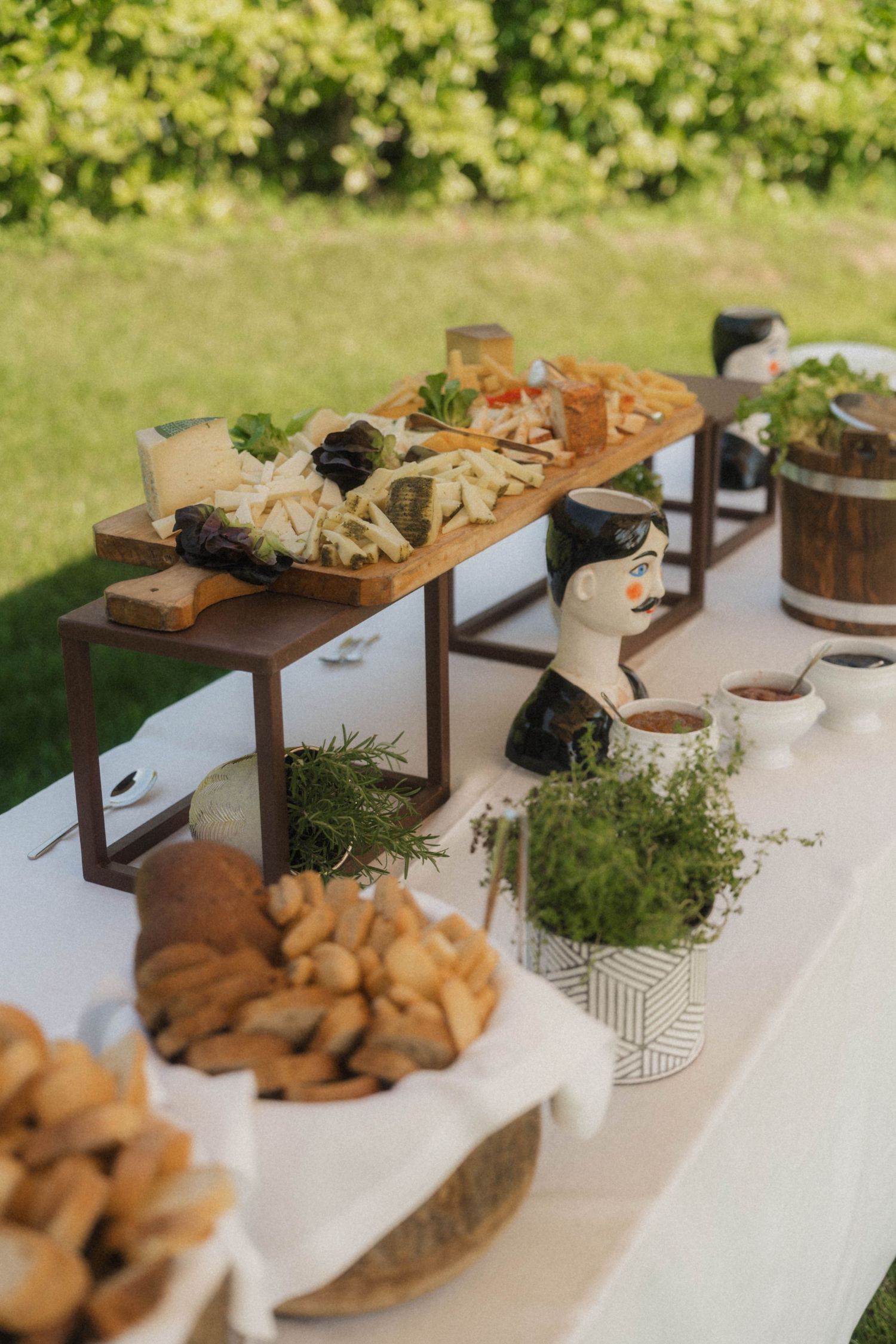 Elegant outdoor wedding reception grazing table with charcuterie, fresh herbs, bread, and gourmet cheeses on tiered wooden stands.