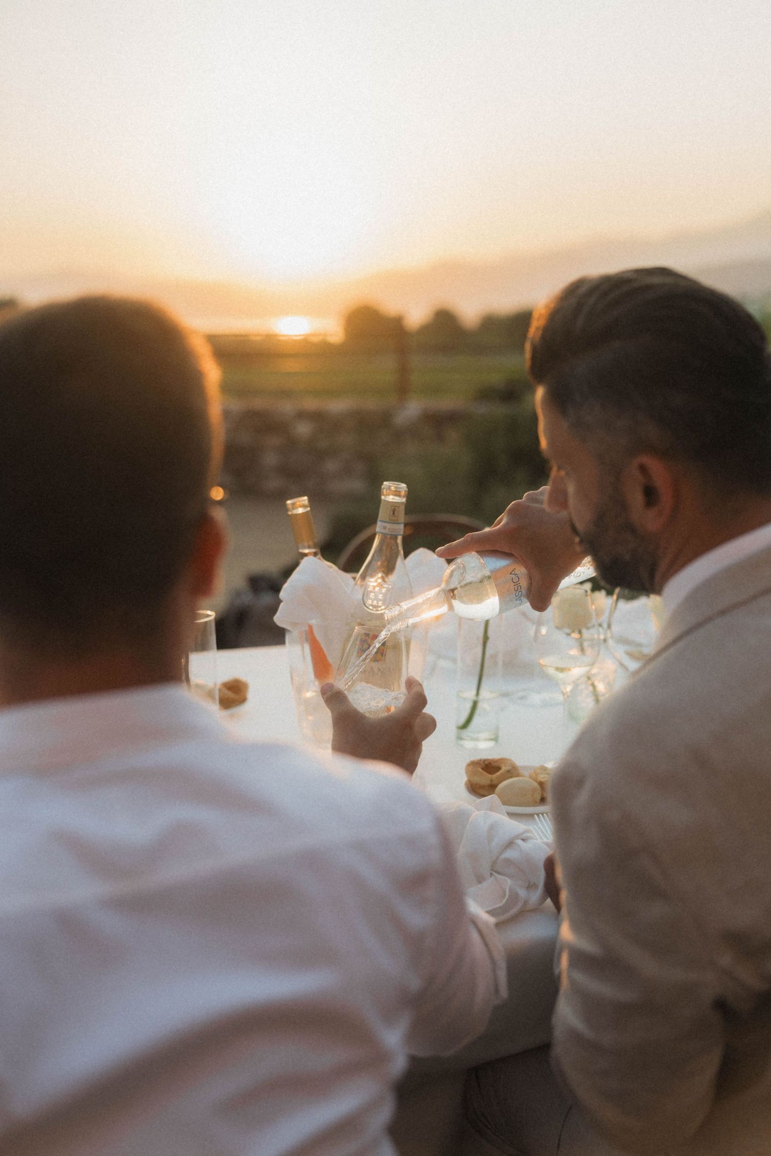 Two men in white shirts clink bottles at an outdoor sunset dinner party.