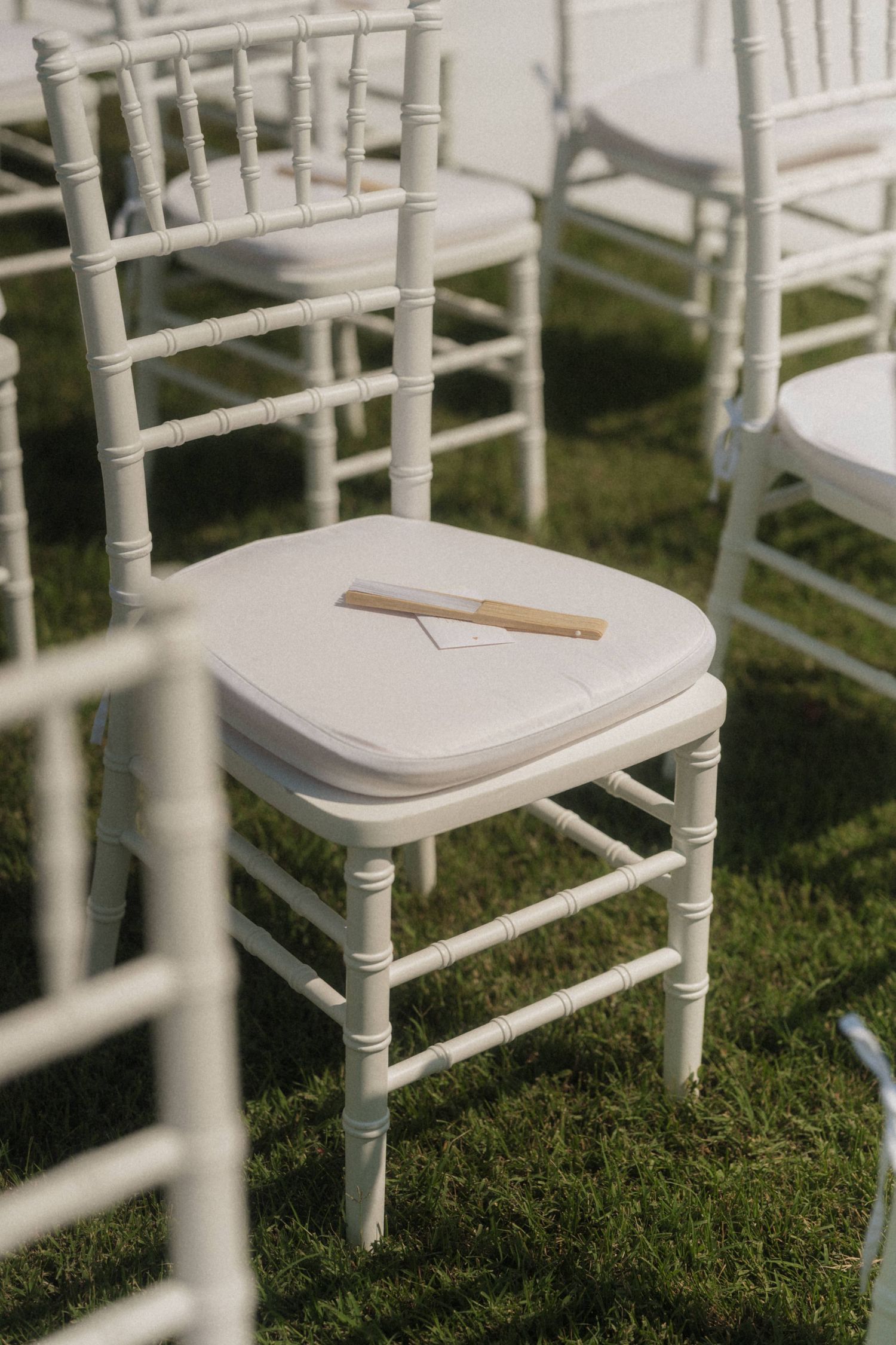 White Chiavari chair with wedding program and fan on grass at outdoor ceremony.