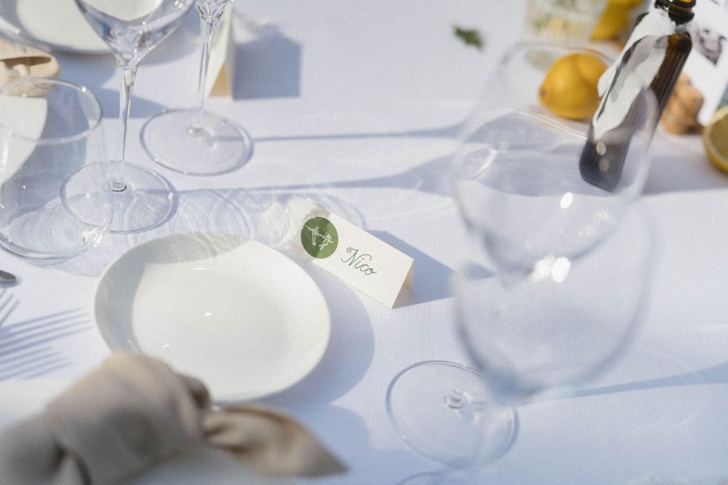 Elegant wedding table setting with white plates, crystal glasses, place cards, and lemons on a white linen tablecloth.