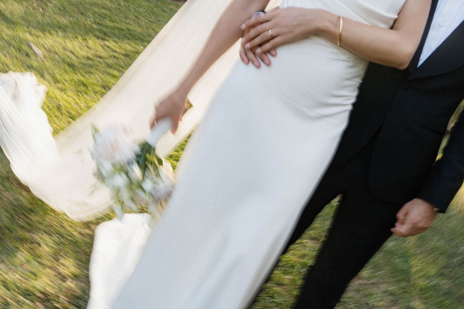 Bride in white gown holding bouquet walks arm in arm with groom in black suit on green grass.