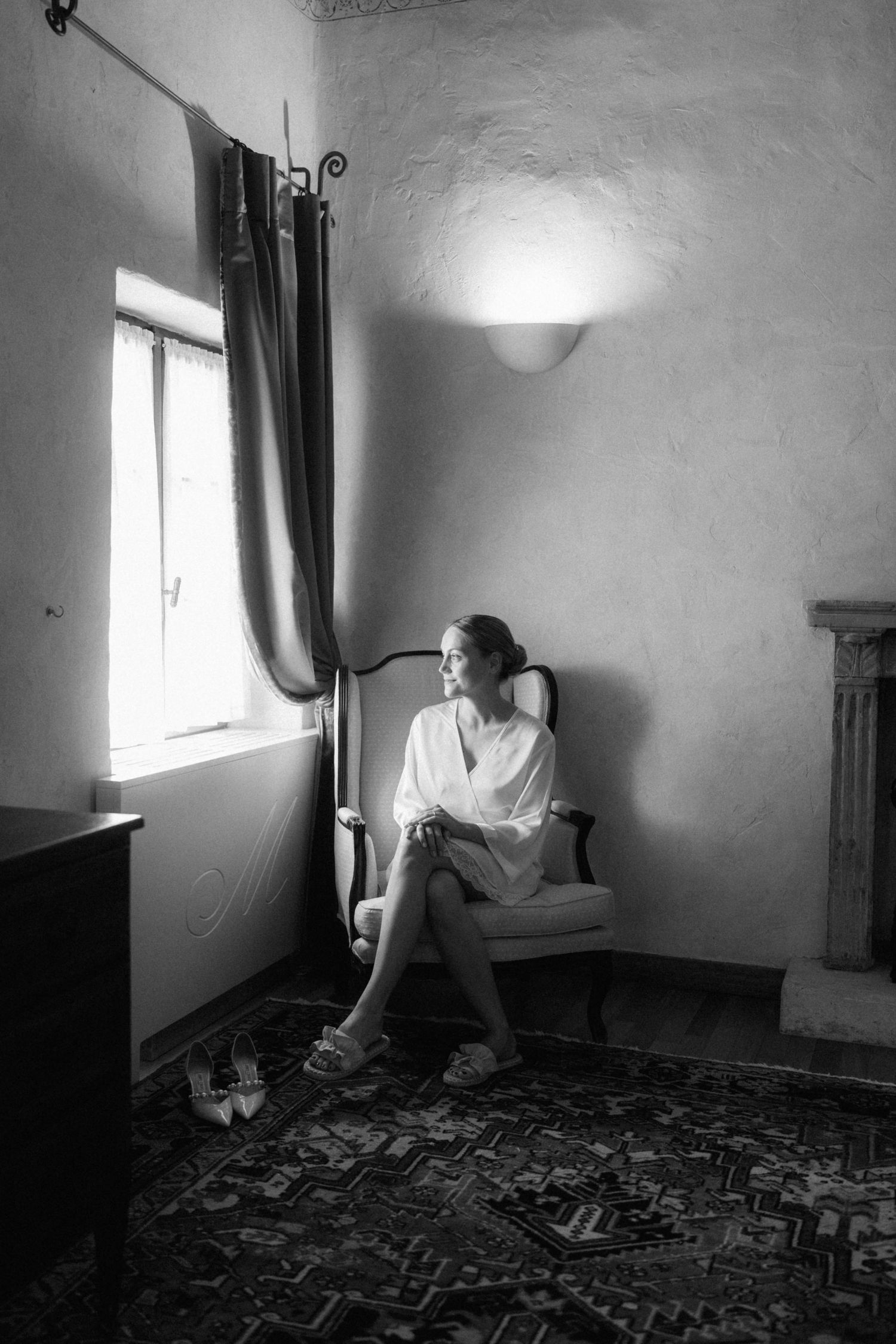 Bride in white robe sits pensively by window in rustic room, black and white wedding preparation photo.