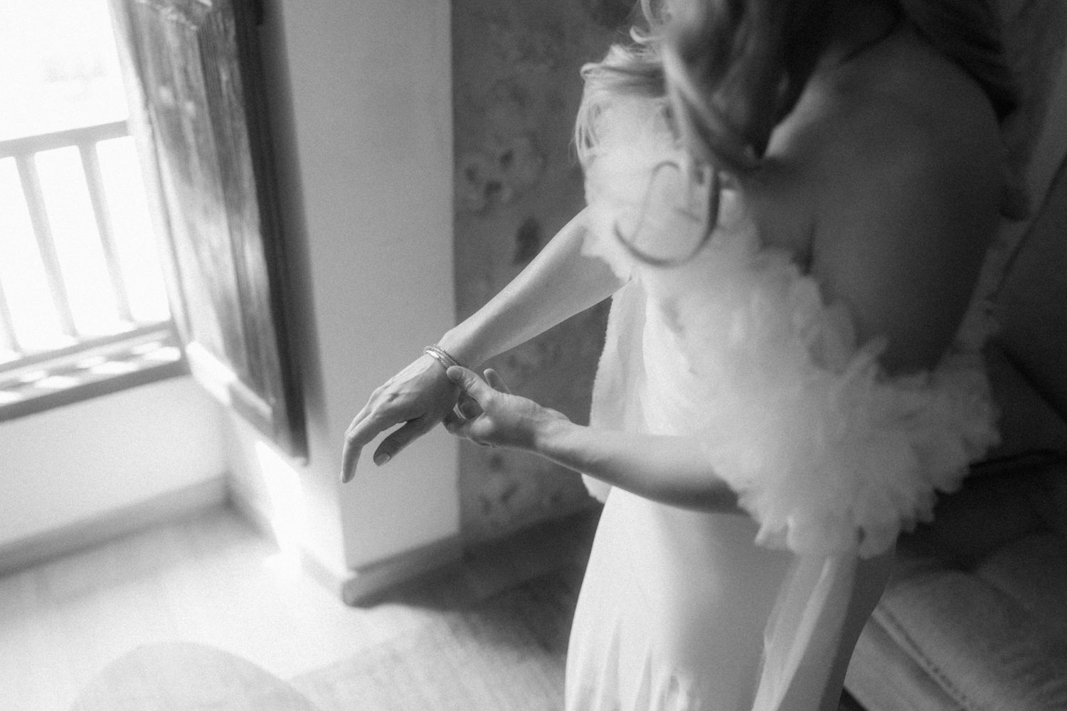 Black and white photo of bride being helped into wedding dress by bridesmaid near sunlit window.
