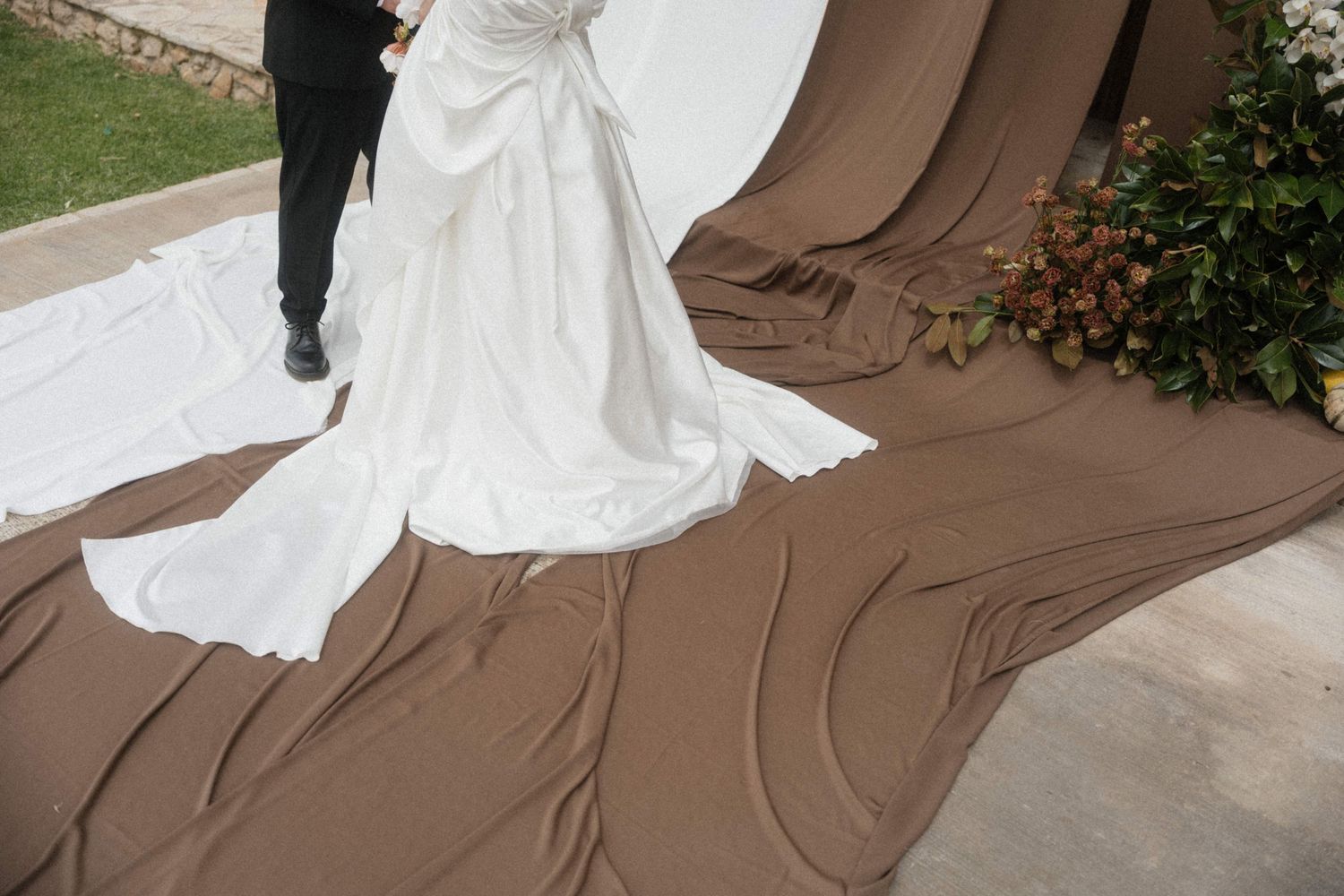 Close-up of bride's white wedding dress train spread over brown fabric aisle runner outdoors.