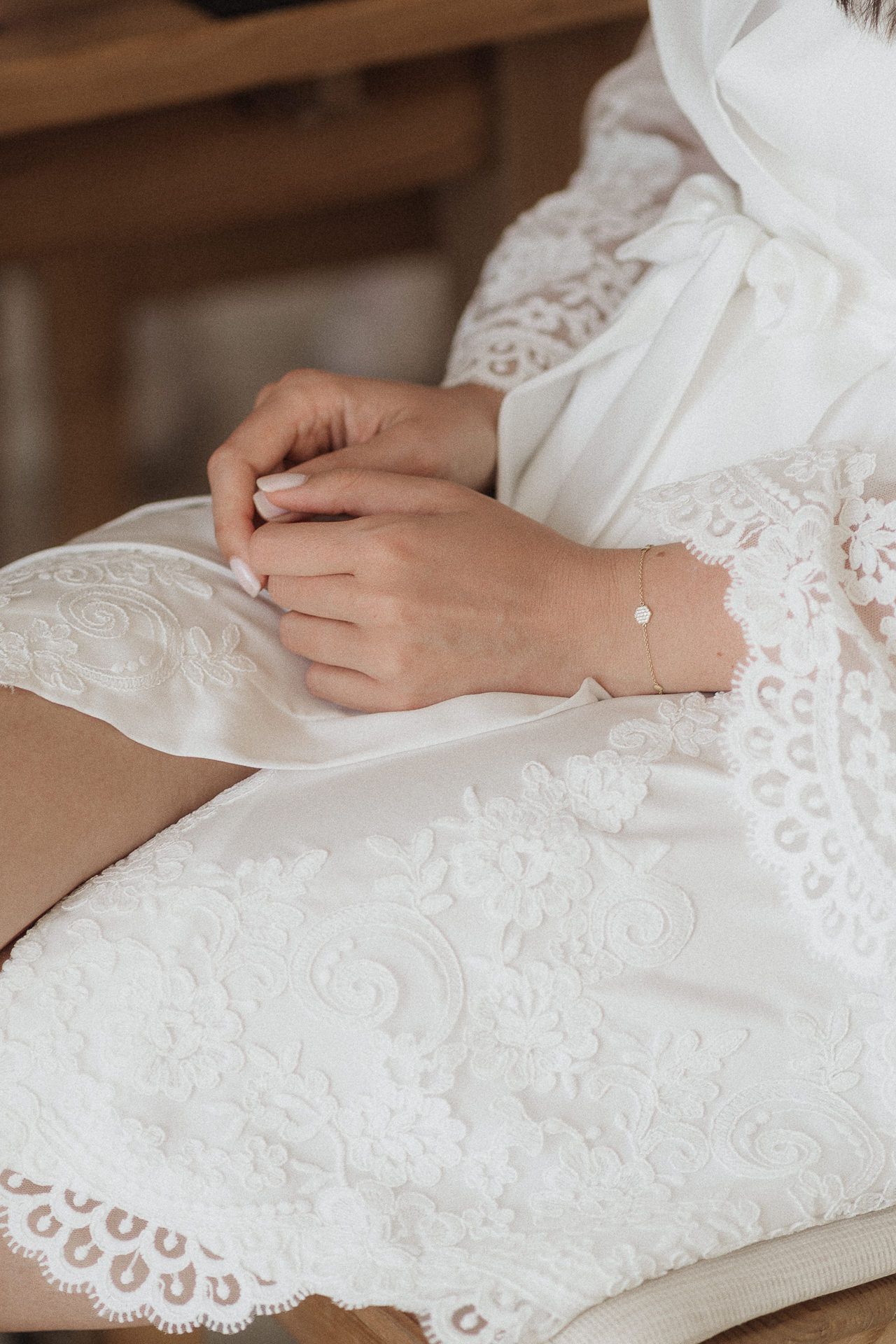 Close-up of a bride in white lace robe with delicate gold bracelet, hands folded gracefully in her lap.