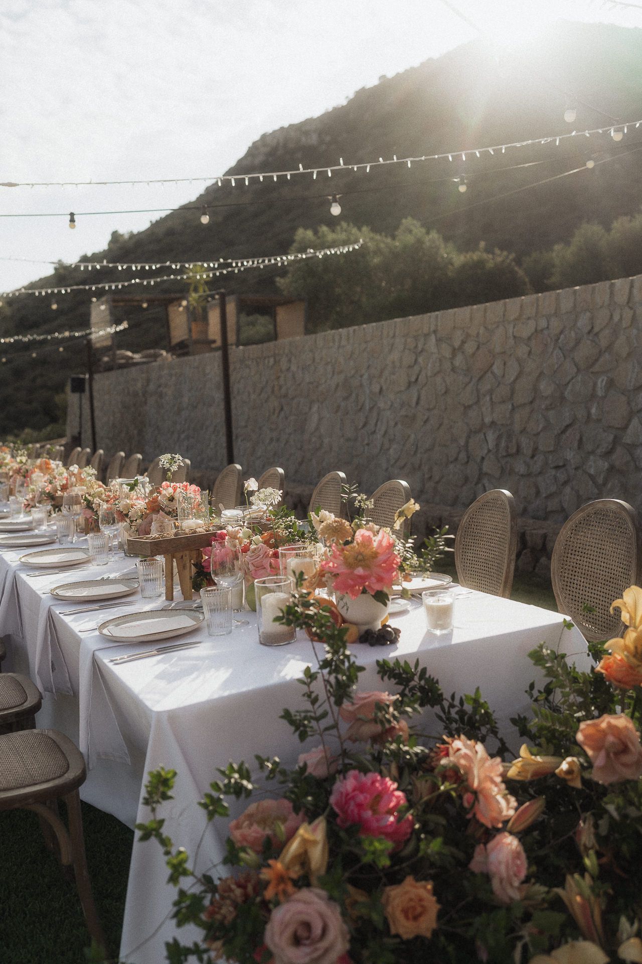Elegant outdoor wedding reception with long white table, pink floral arrangements, string lights, and stone wall backdrop.