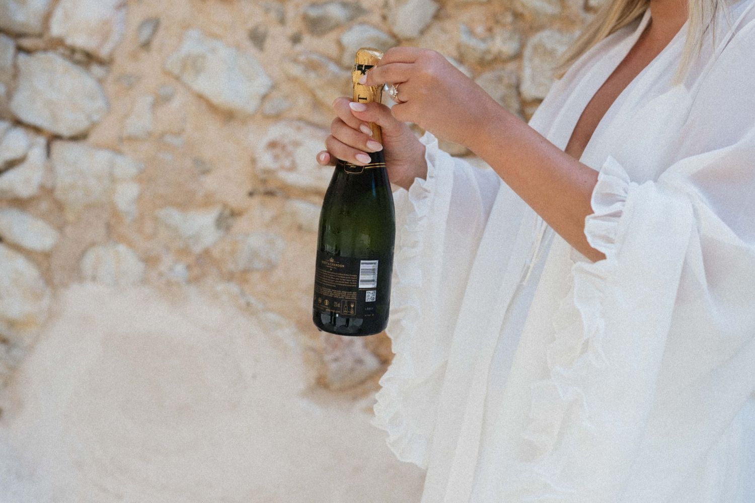 Bride in white dress holding a champagne bottle against a rustic stone wall.