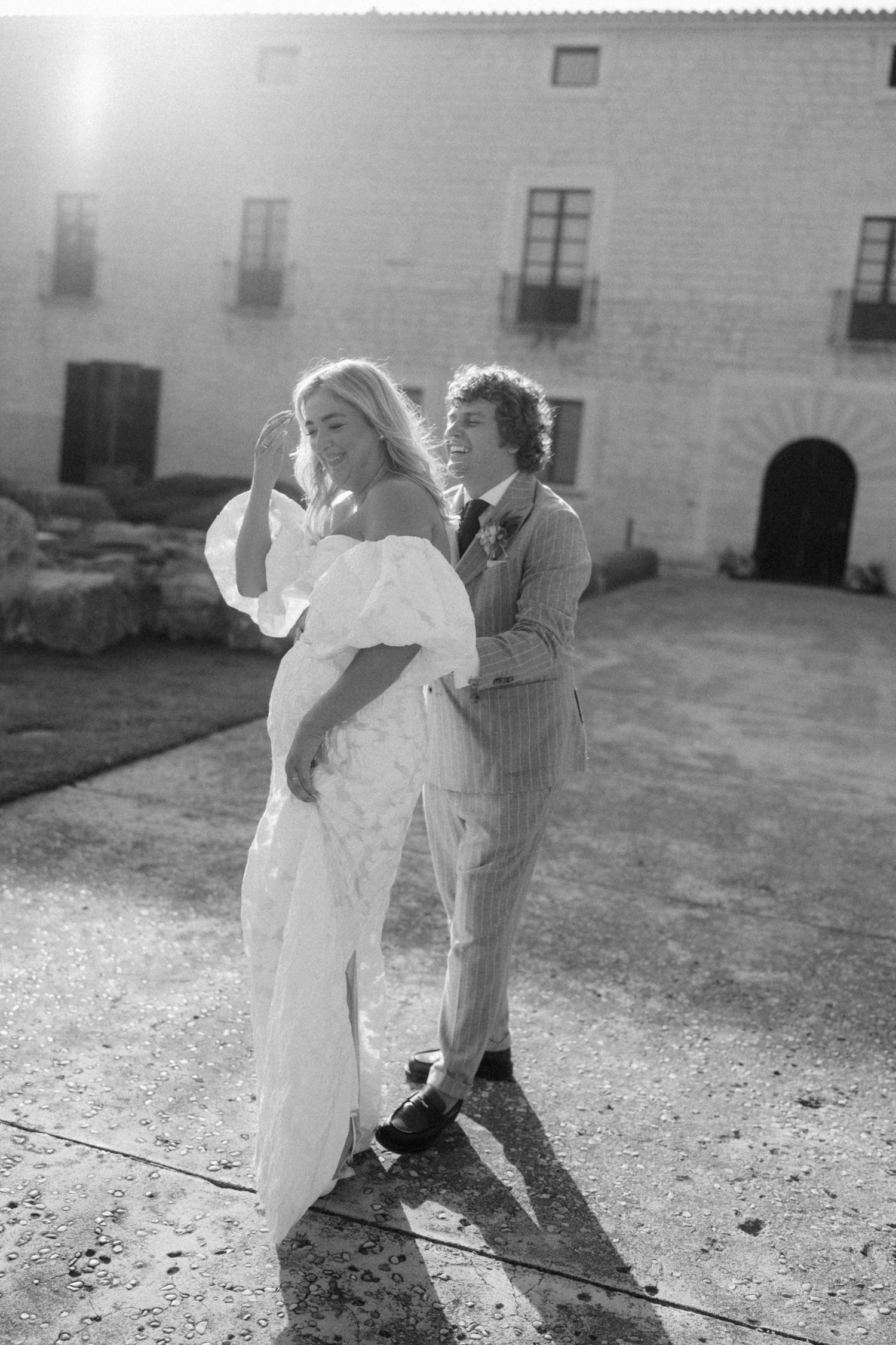 Black and white photo of bride and groom dancing in sunlight outside a Spanish stone estate.