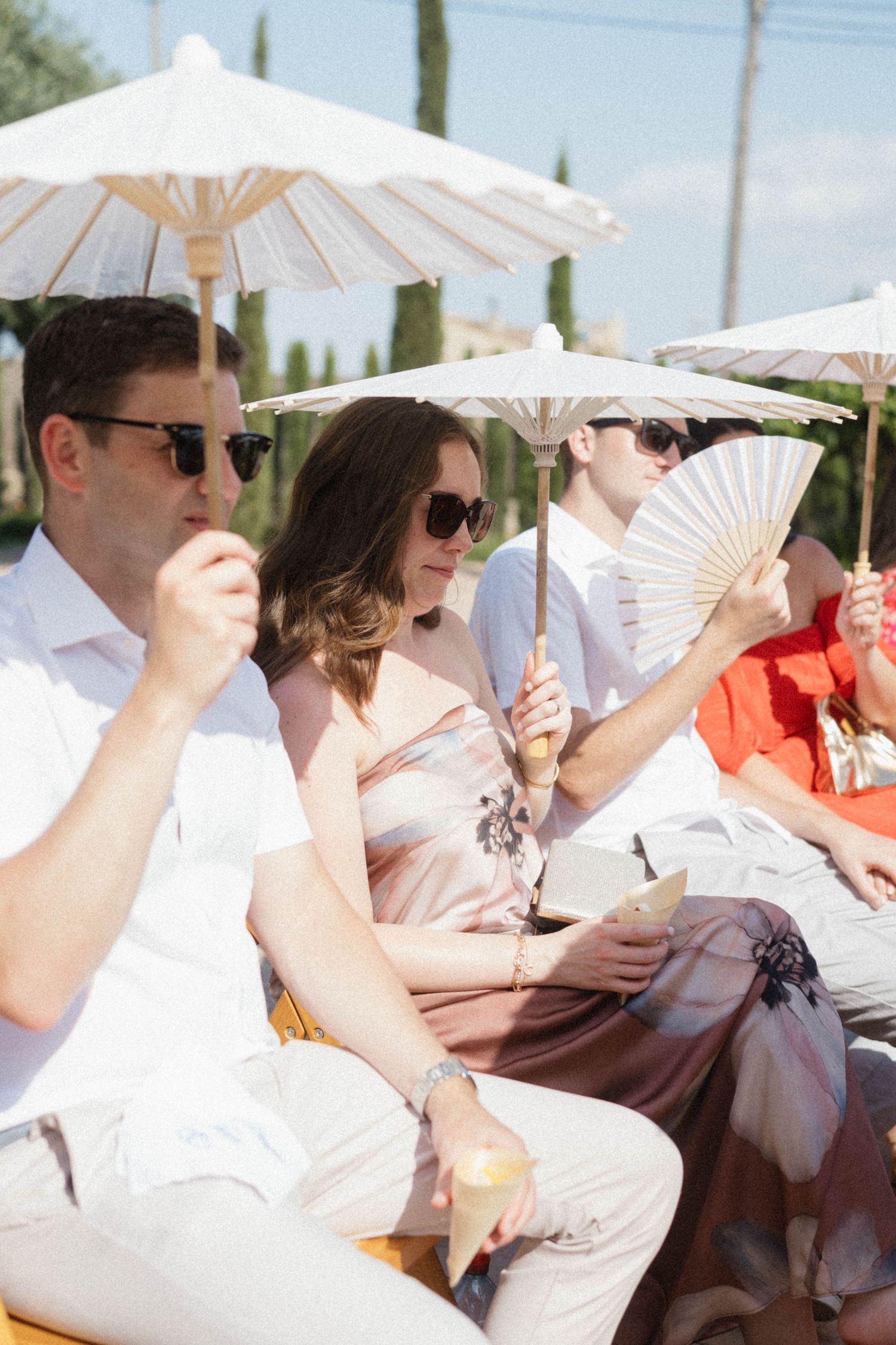 Guests at an outdoor wedding use parasols and hand fans to stay cool in the summer heat.