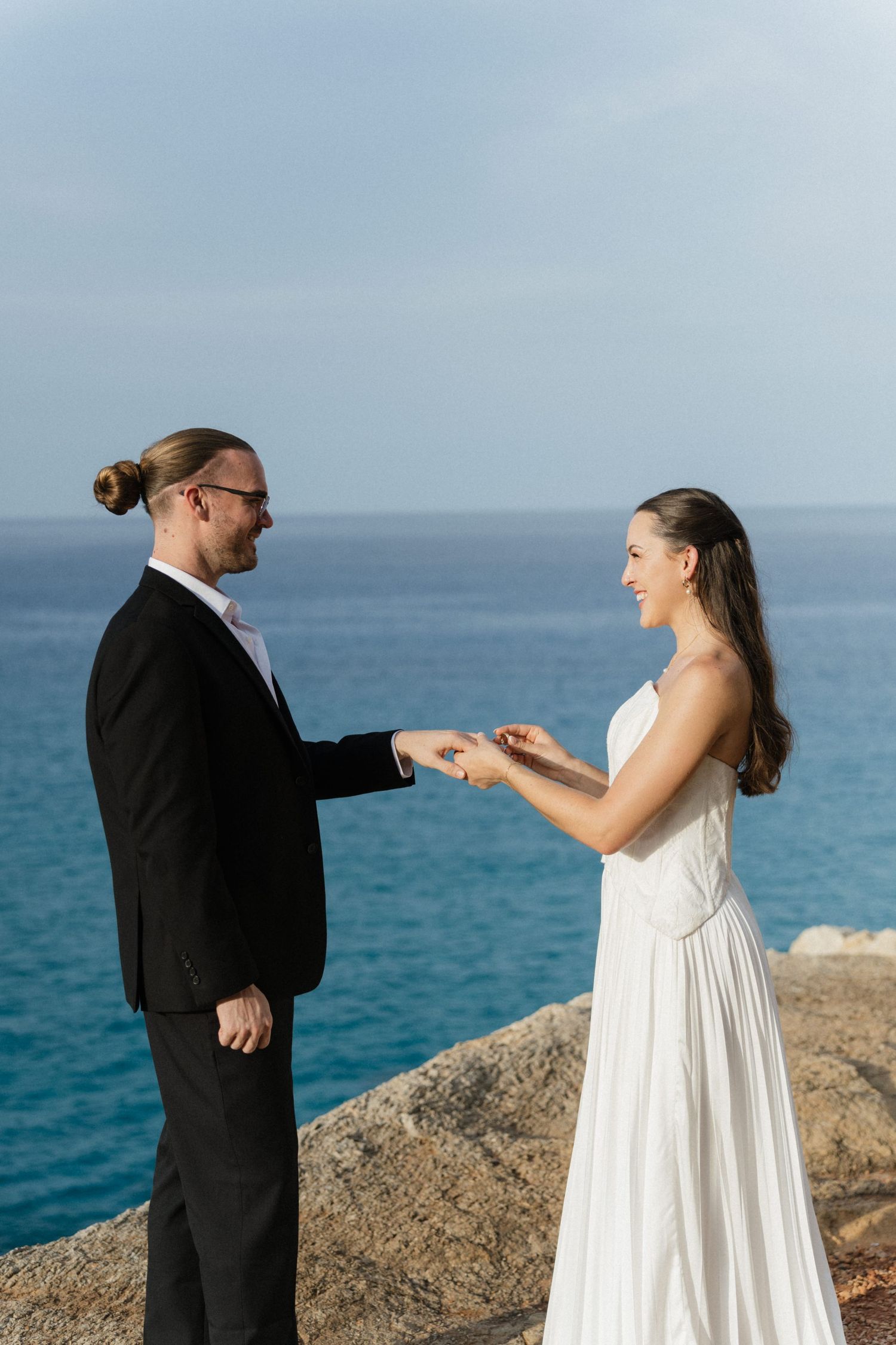 Groom with a man bun and glasses exchanges rings with his smiling bride on a sunny cliff above the blue sea.
