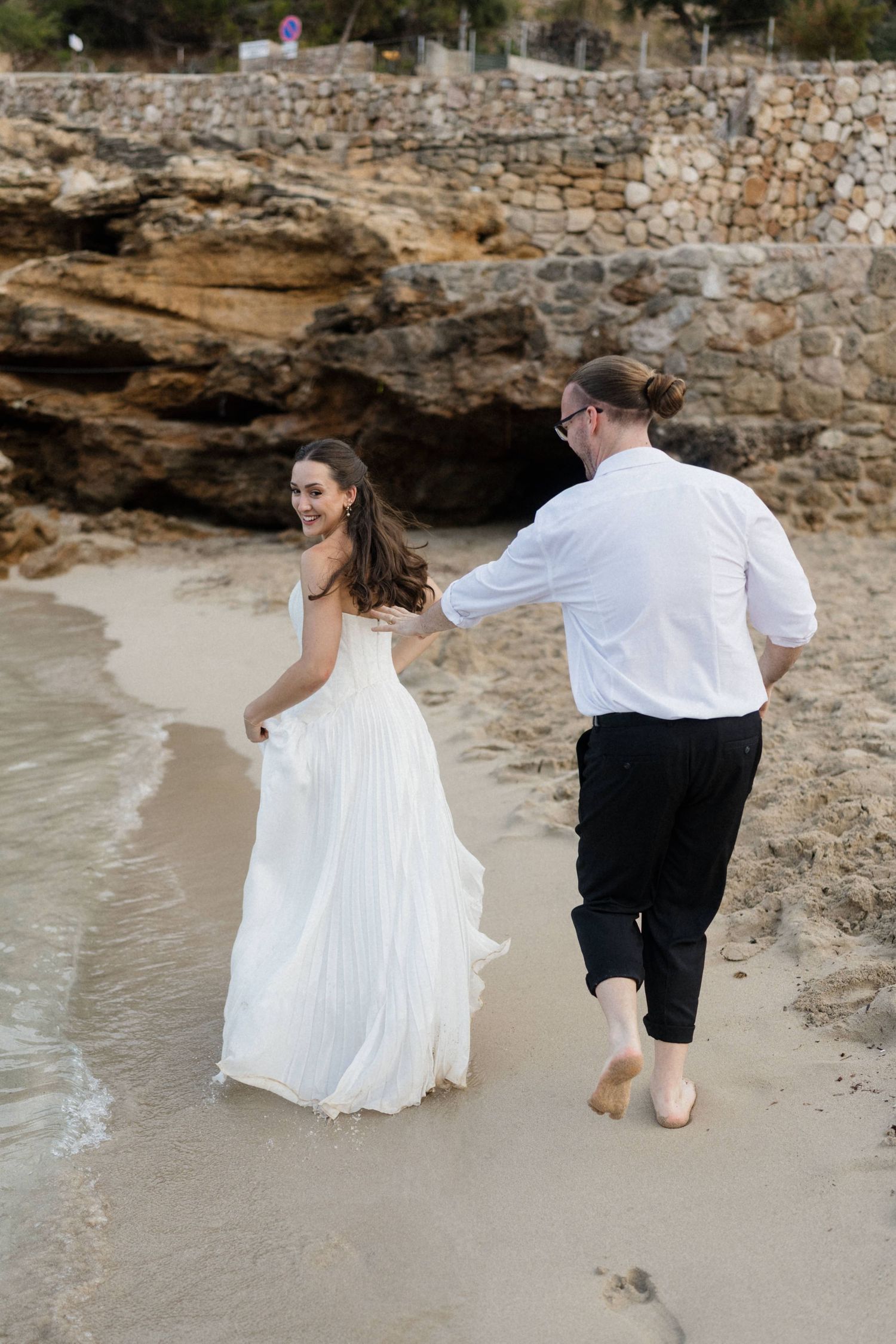 Bride in a white gown looks back smiling as she holds hands with a barefoot groom running along a rocky beach shoreline.
