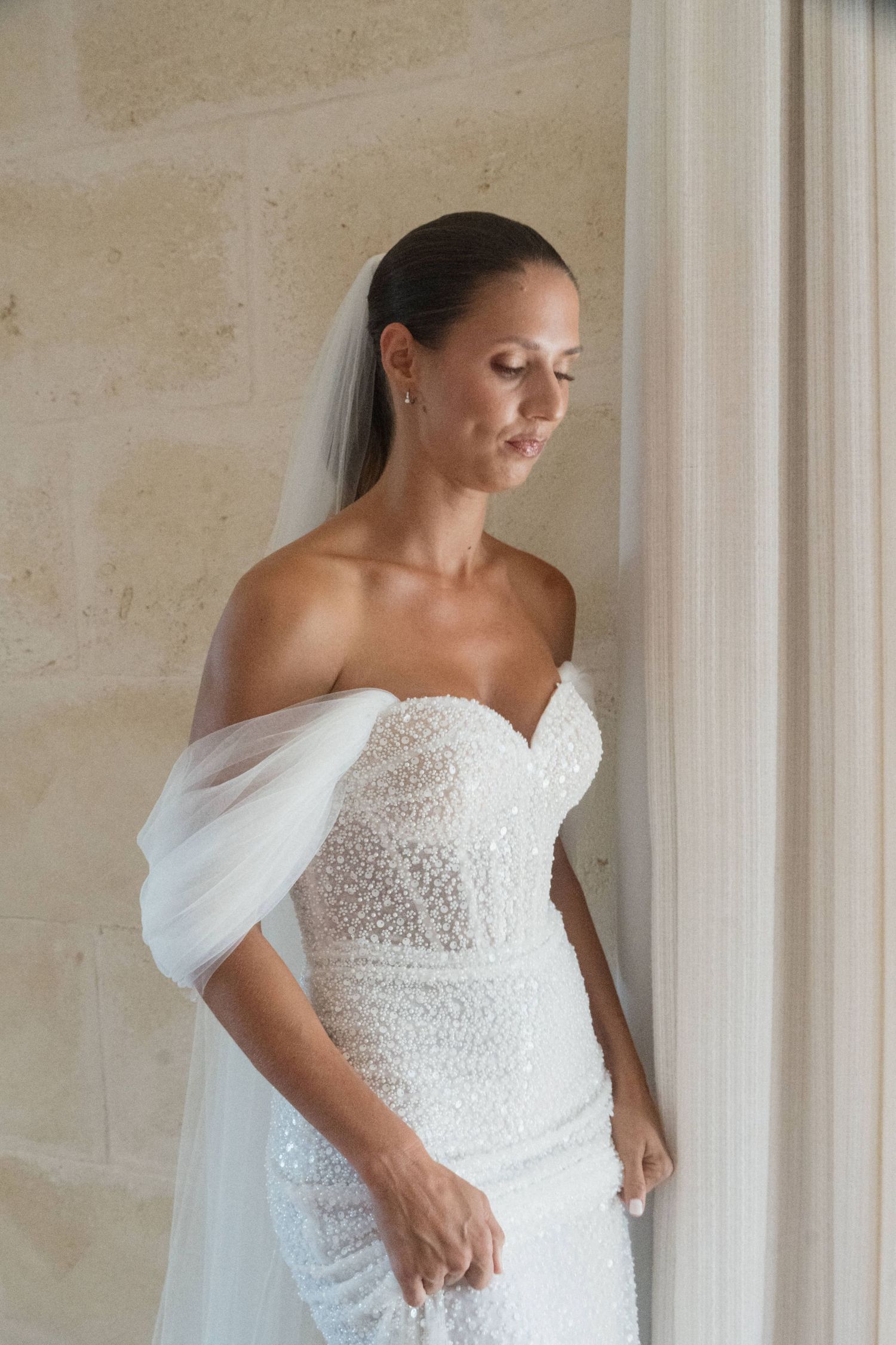 Bride in an off-the-shoulder sequined wedding gown and long veil looks down while standing beside a limestone wall and curtain.