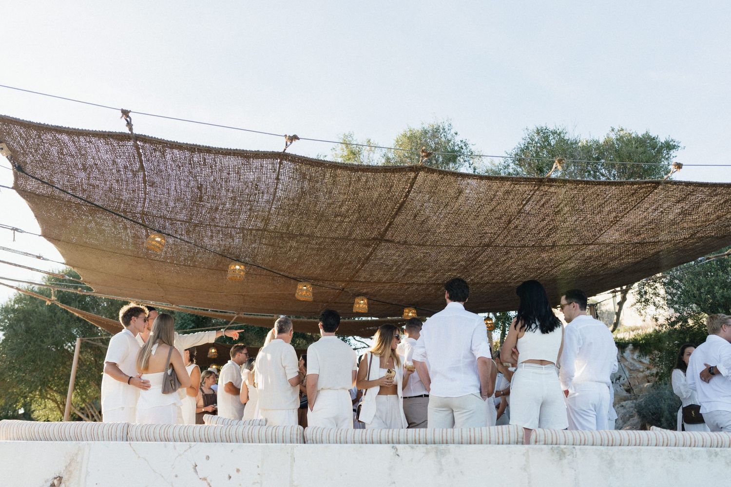 Guests dressed in white gather beneath a rustic woven reed canopy at an outdoor celebration surrounded by trees.