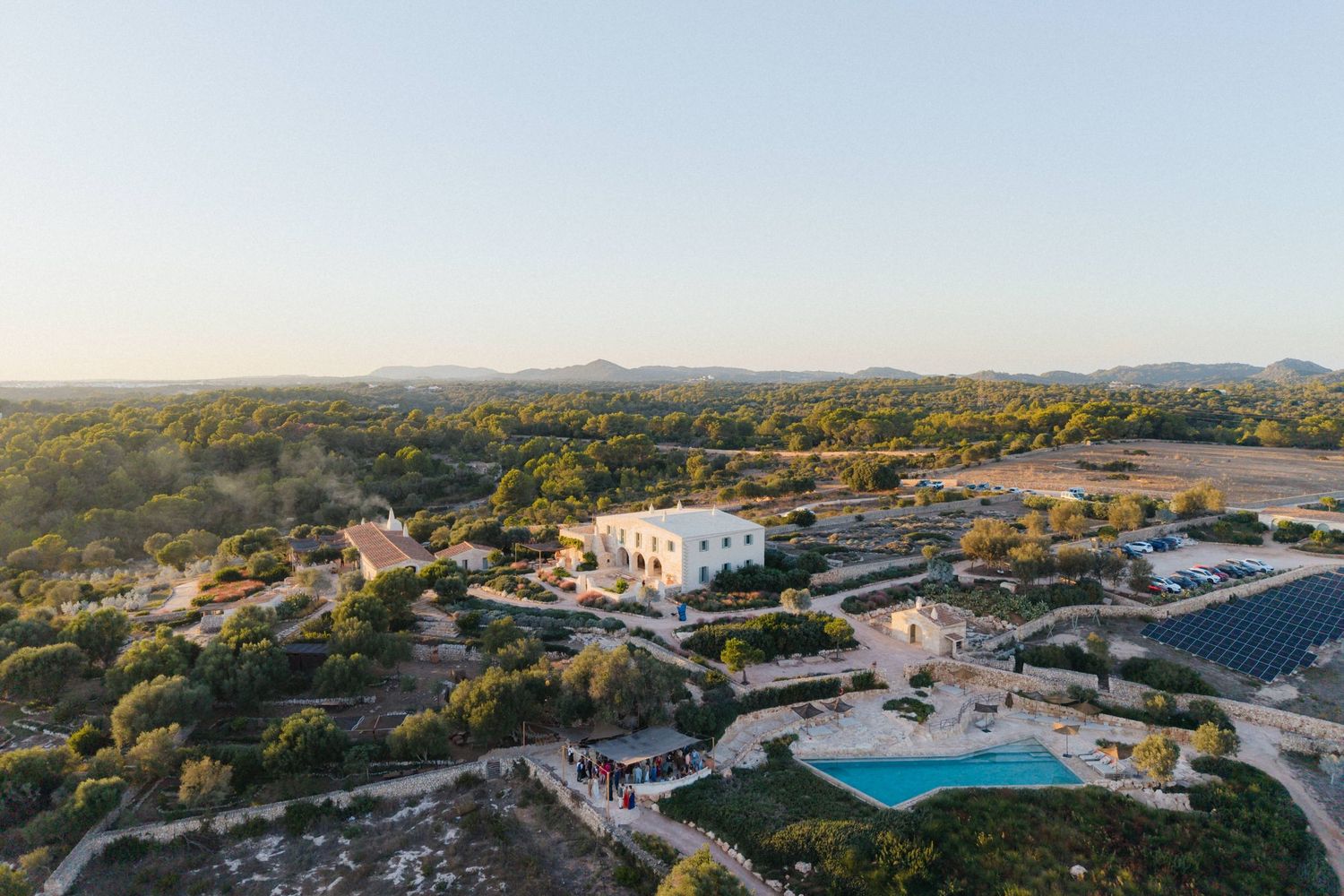 Aerial view of a whitewashed countryside wedding venue with a swimming pool surrounded by trees and gardens.