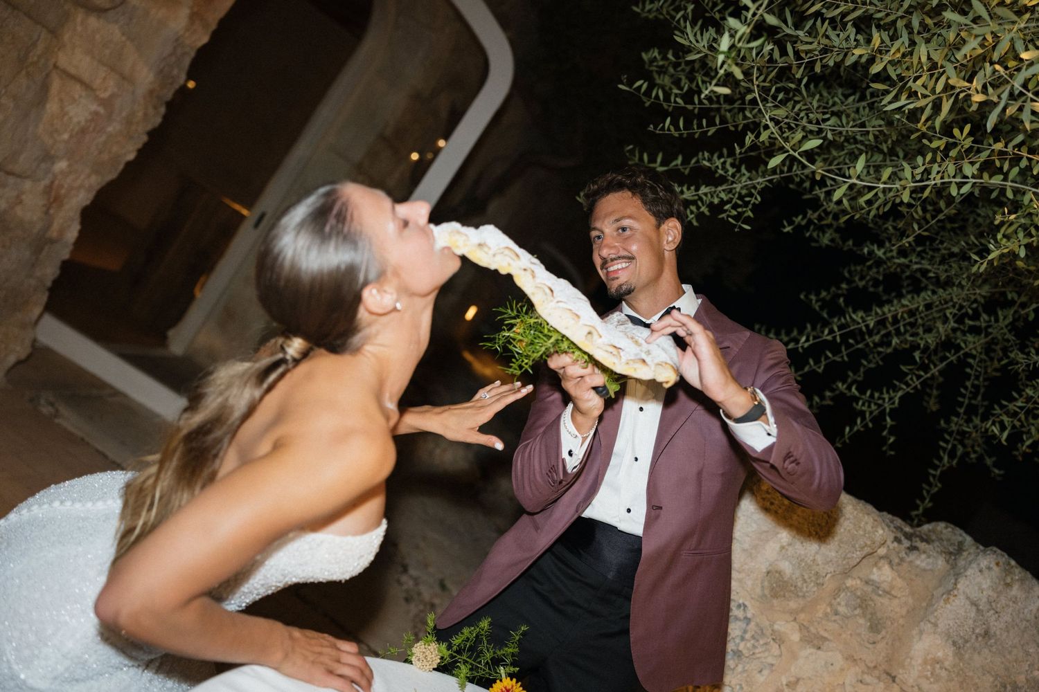 Bride in a white gown playfully takes a bite from a decorated treat held by her laughing groom in a burgundy suit at night.