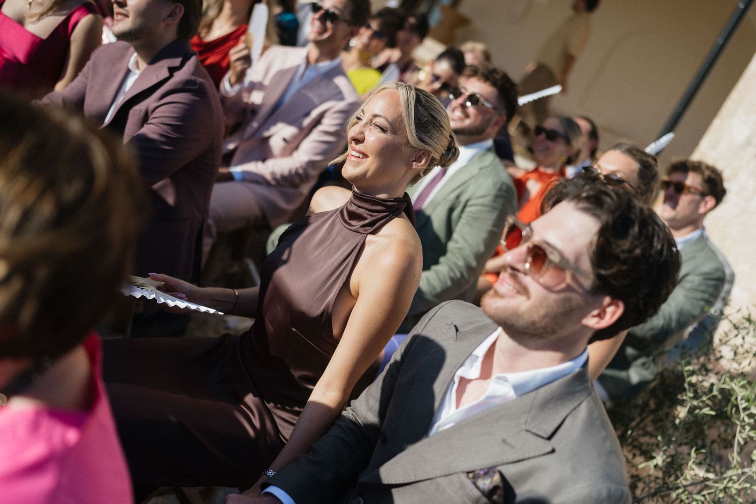 Wedding guests in stylish outfits and sunglasses sit outdoors during a ceremony, smiling in the warm sunlight.
