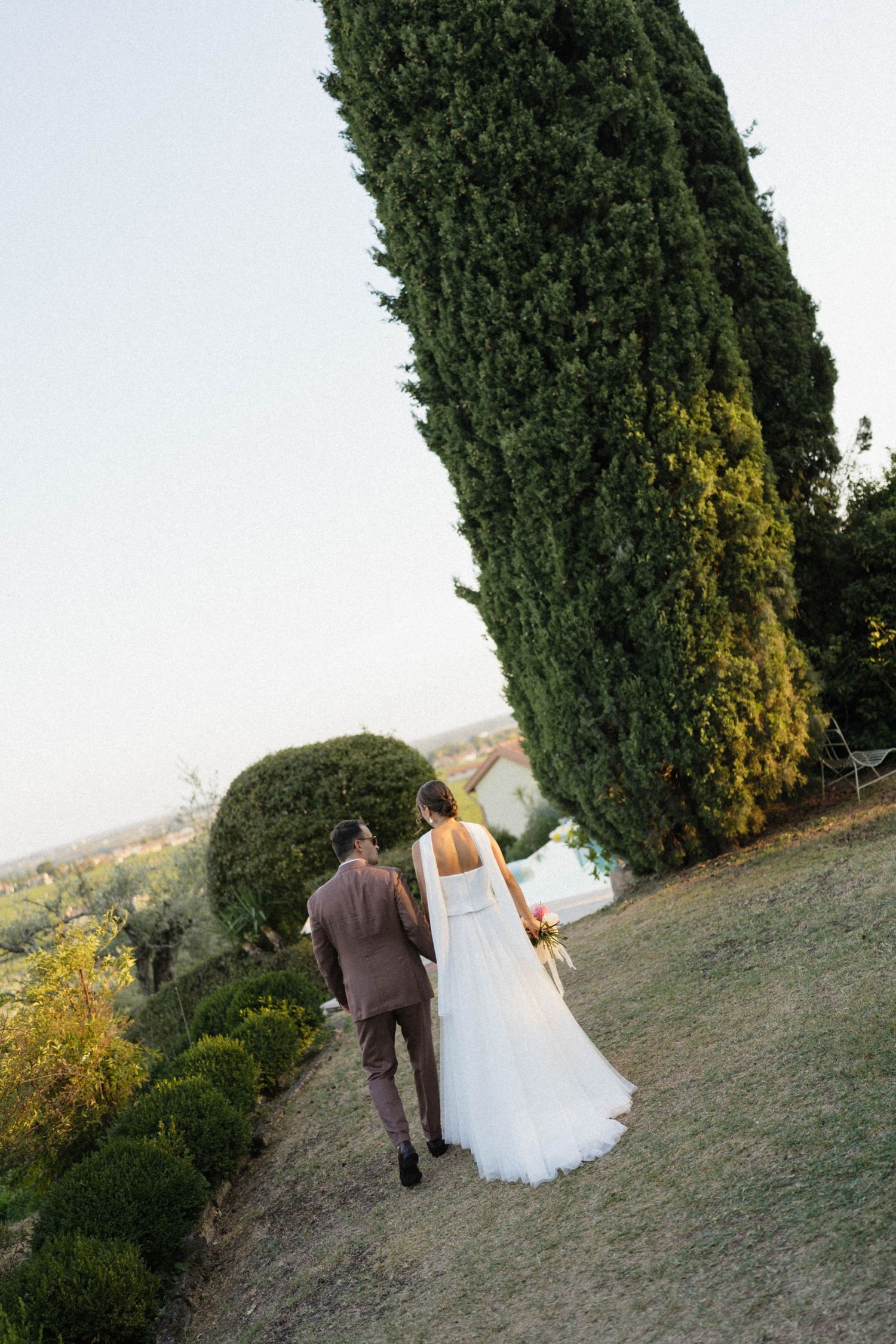 Bride and groom walking hand in hand through lush garden grounds with tall cypress tree on their wedding day.