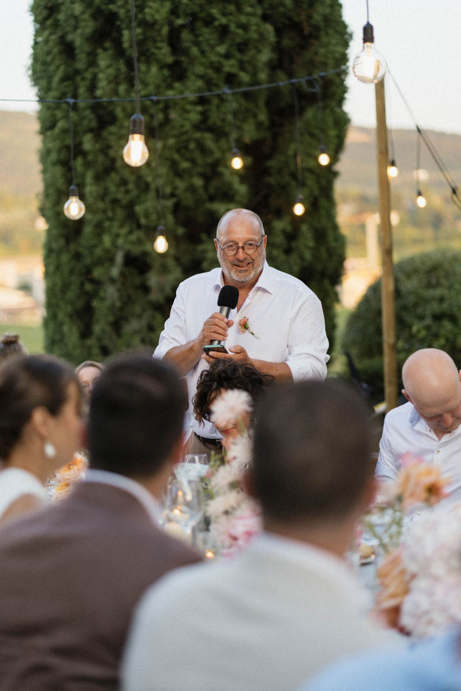 Older man in white shirt giving a speech with microphone at outdoor wedding reception with string lights.