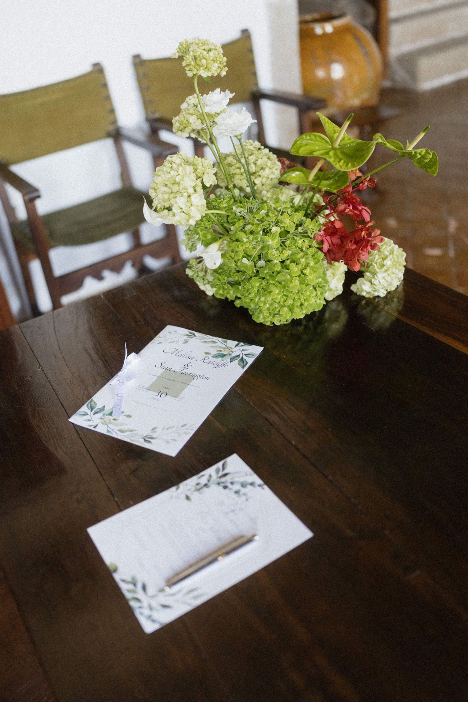 Wedding programs and floral centerpiece with white hydrangeas on a dark wooden table indoors.