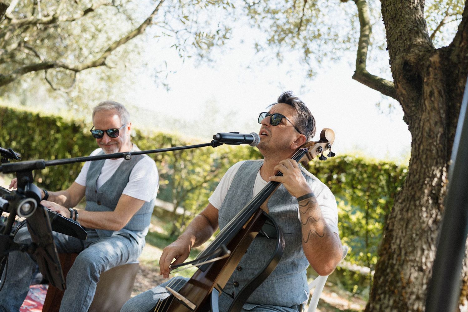 Two men playing guitar and cello outdoors under a tree on a sunny day.