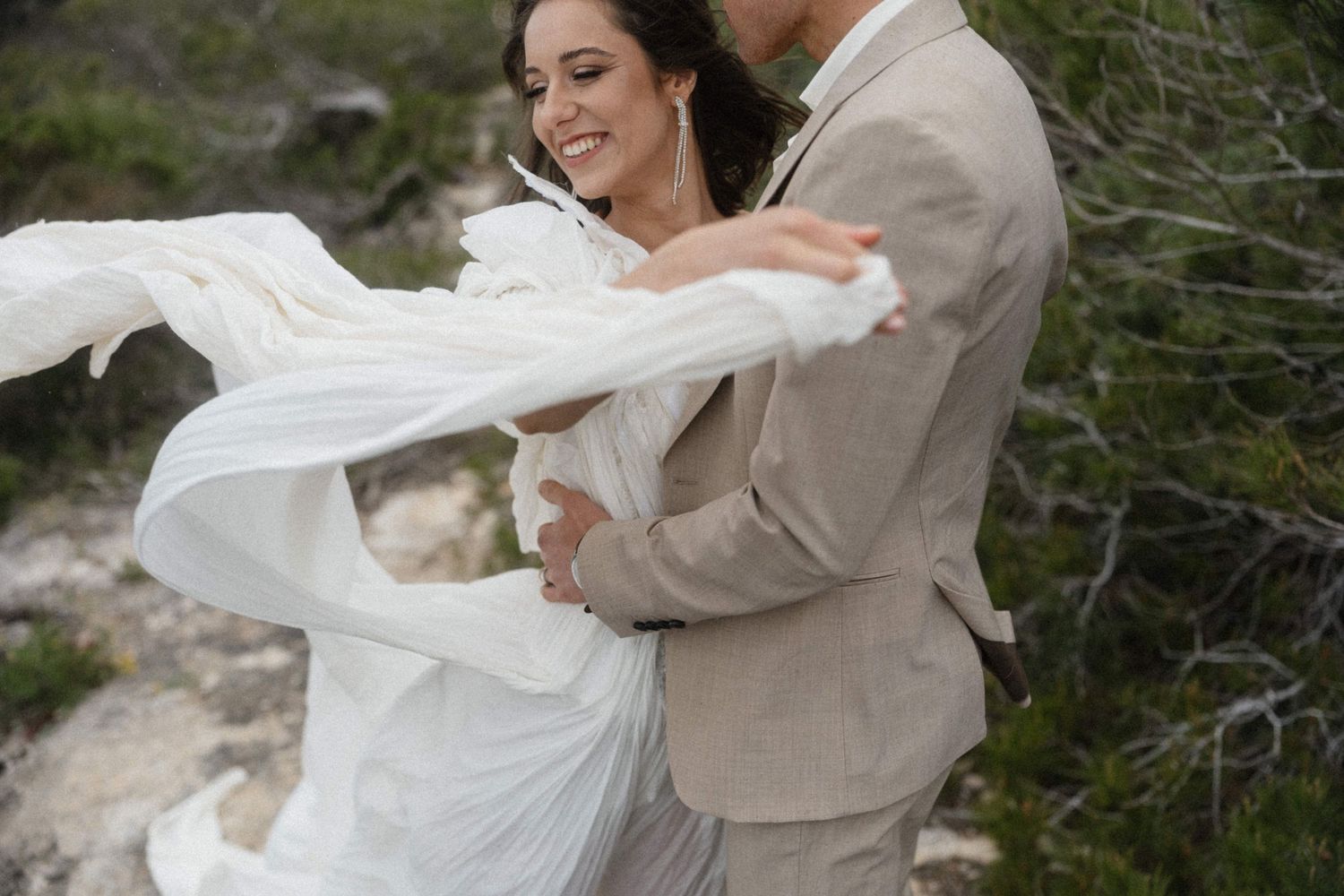 Bride in flowing white dress laughs joyfully as groom in tan suit embraces her outdoors on rocky terrain.