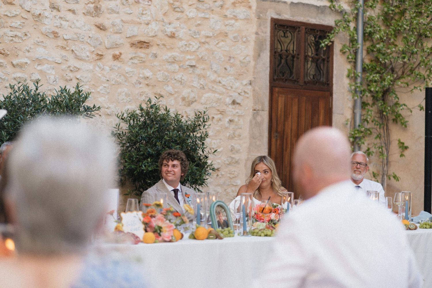 Bride and groom seated at outdoor wedding reception table at a rustic stone venue with colorful floral arrangements.