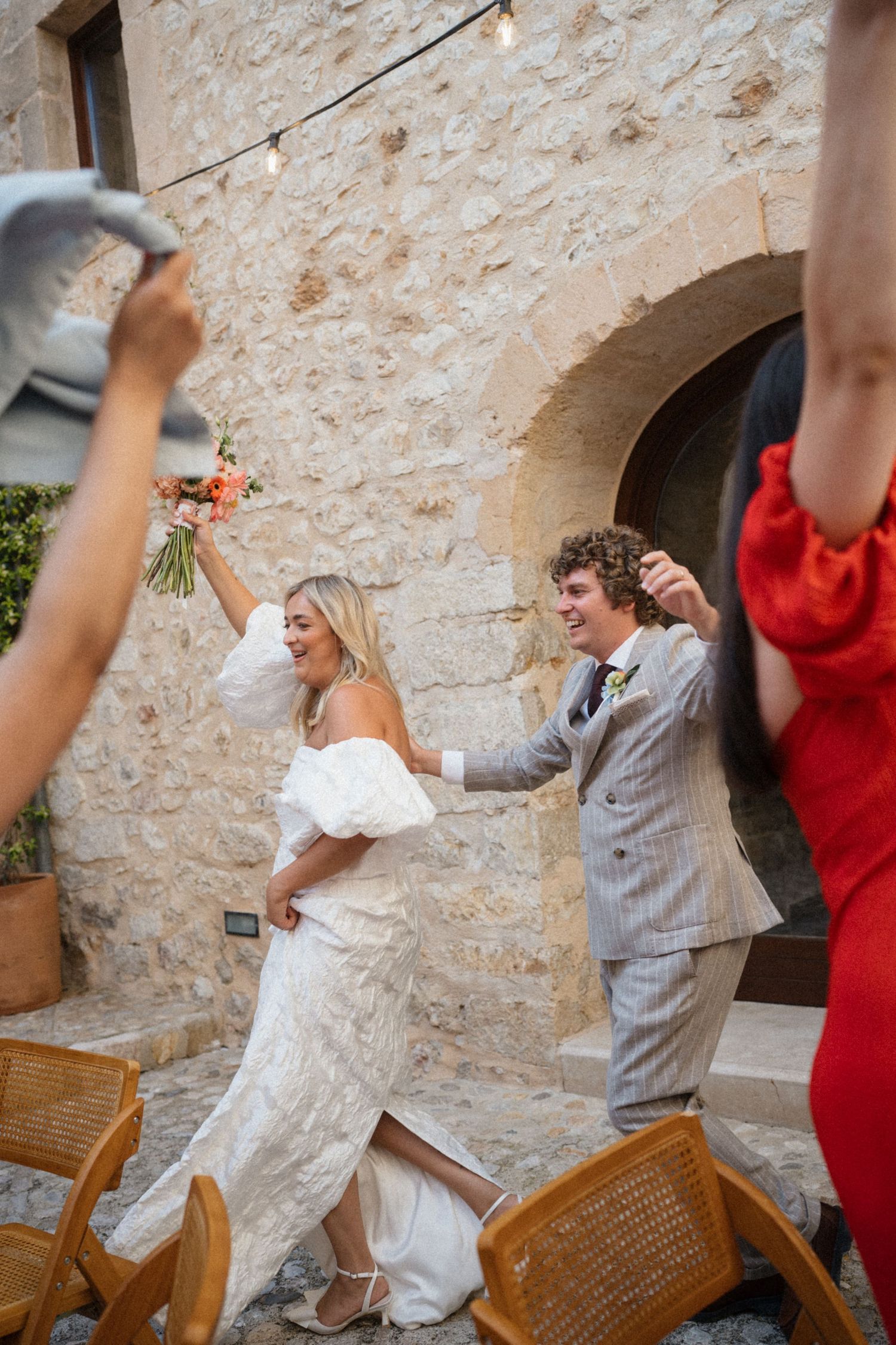 Joyful newlyweds celebrate their wedding ceremony exit at a rustic stone venue, bride holding bouquet aloft.