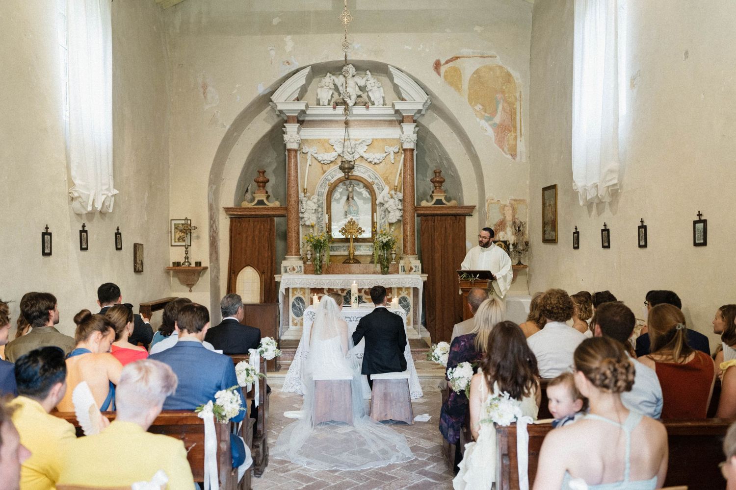 Guests seated in a charming Italian church witness a couple kneeling at the altar during their wedding ceremony.