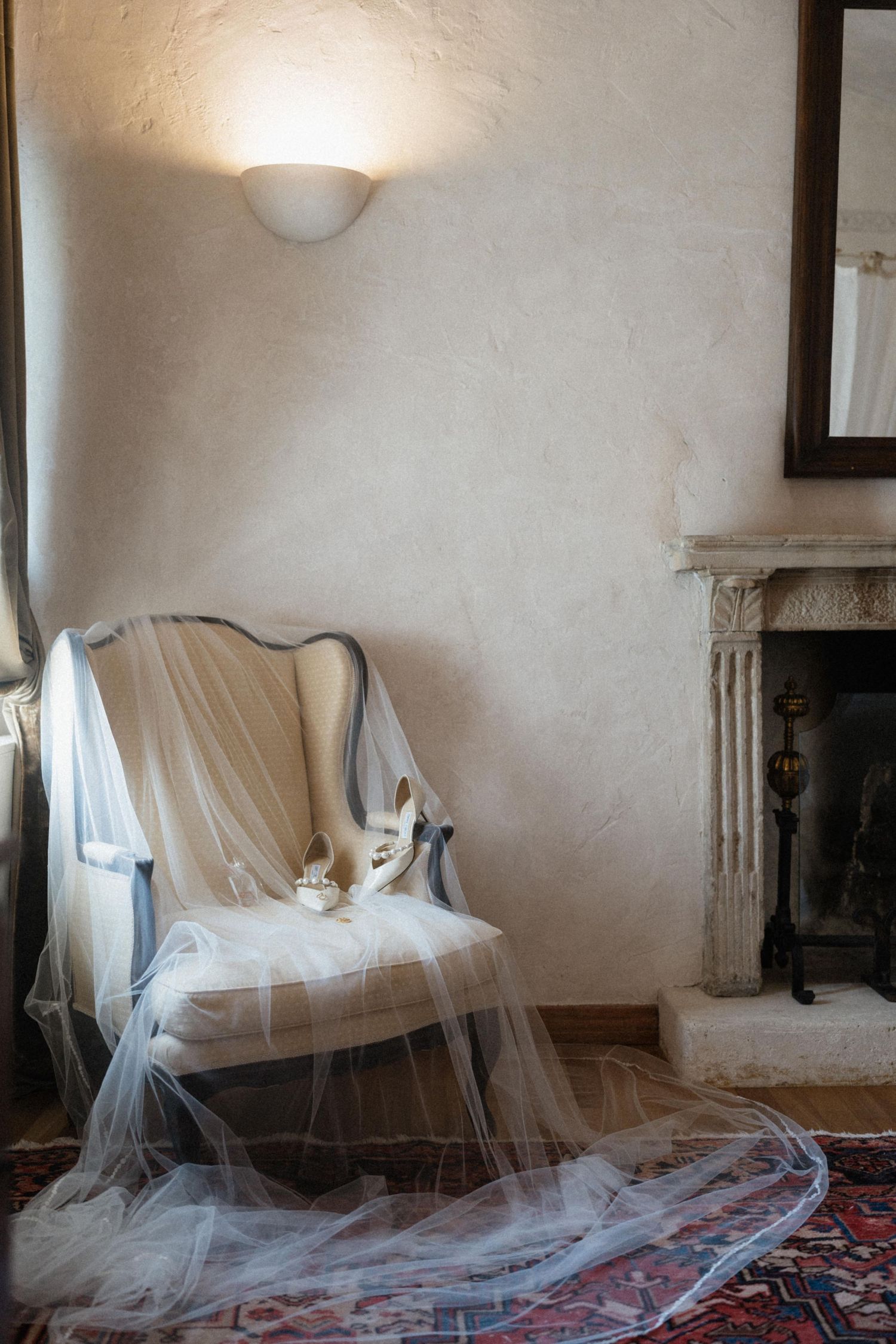 Elegant wedding veil and bridal shoes draped over a vintage blue chair near a classic fireplace.