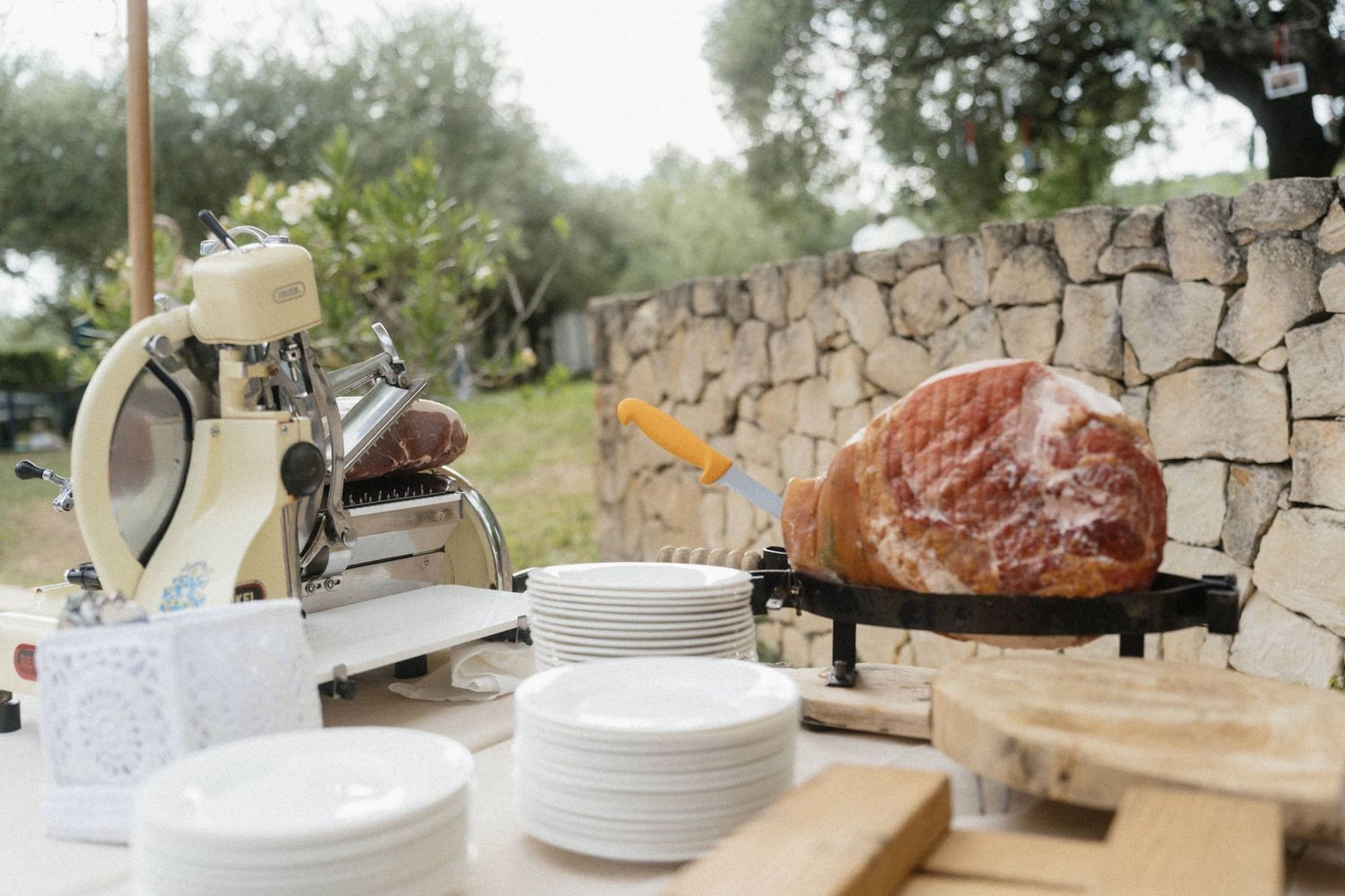 Outdoor charcuterie station with a meat slicer, stacked white plates, and a whole prosciutto on a carving stand.