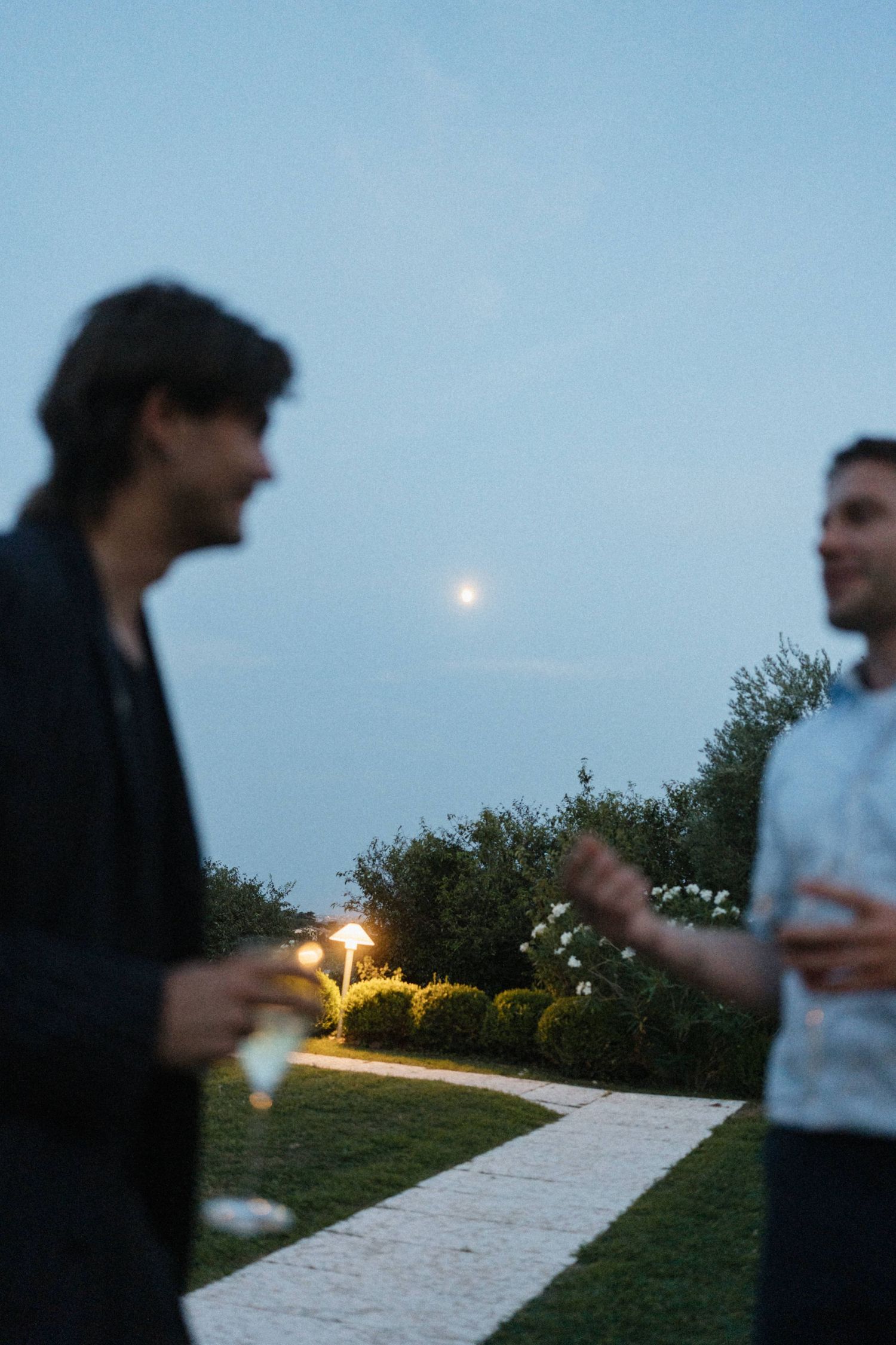 Two men conversing outdoors at dusk with a full moon rising over a manicured garden pathway with soft landscape lighting.