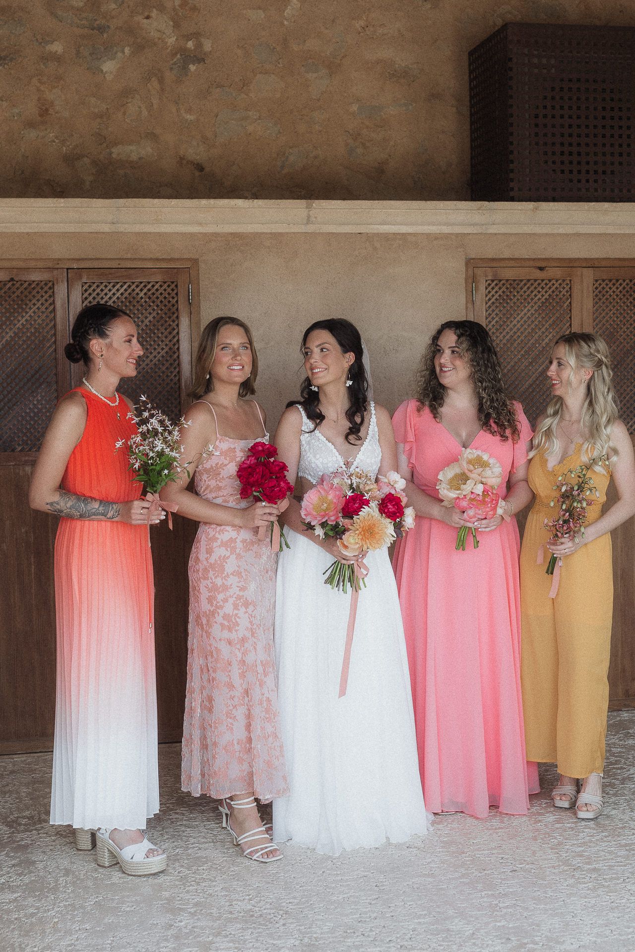 Bride in white gown with four bridesmaids in colorful dresses holding bouquets, posing together at a wedding venue.