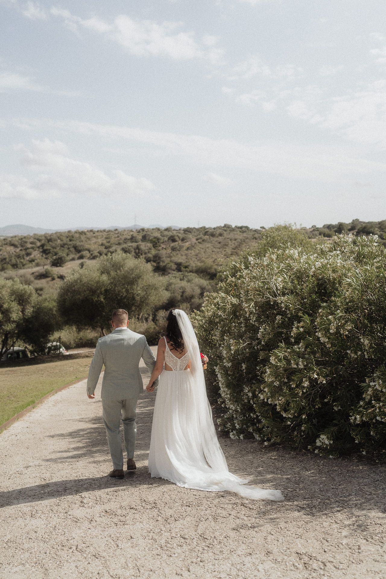 Bride and groom walking hand in hand down a gravel path surrounded by lush greenery on their wedding day.